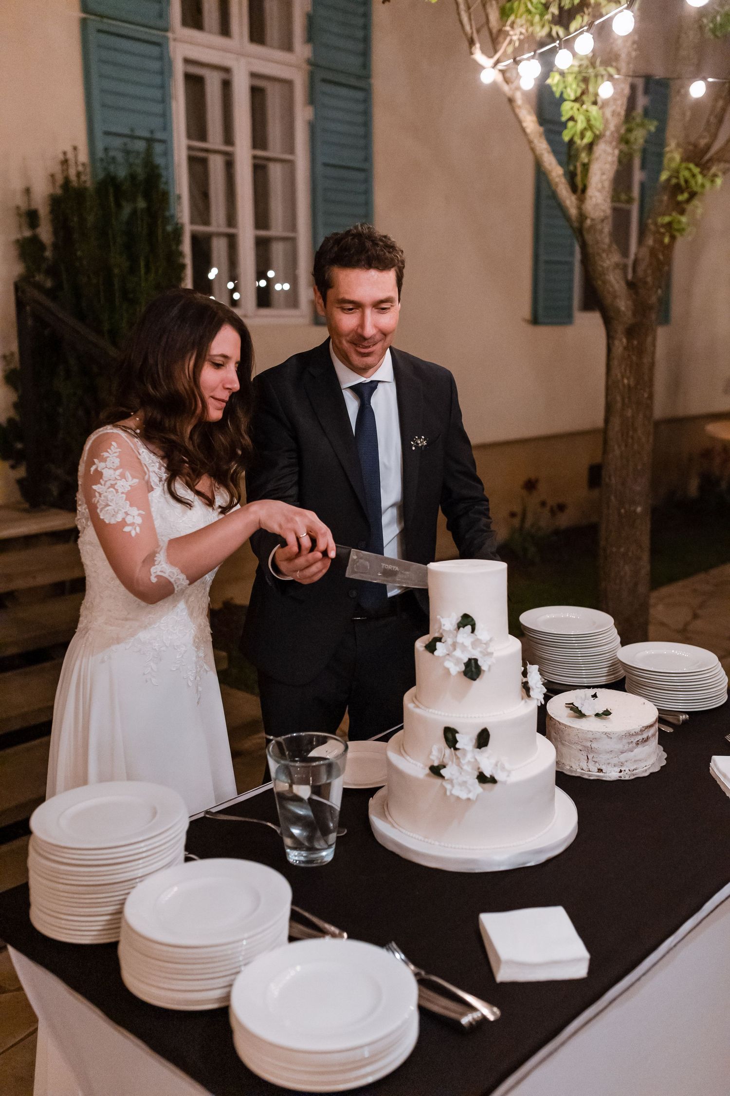 Newlyweds cut their elegant white wedding cake adorned with black flowers at an evening outdoor reception.