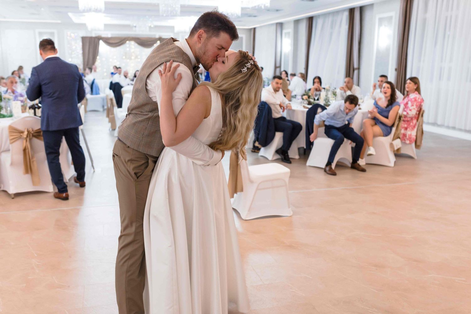 A couple shares their first dance at a wedding reception while guests watch from chairs along the wall.
