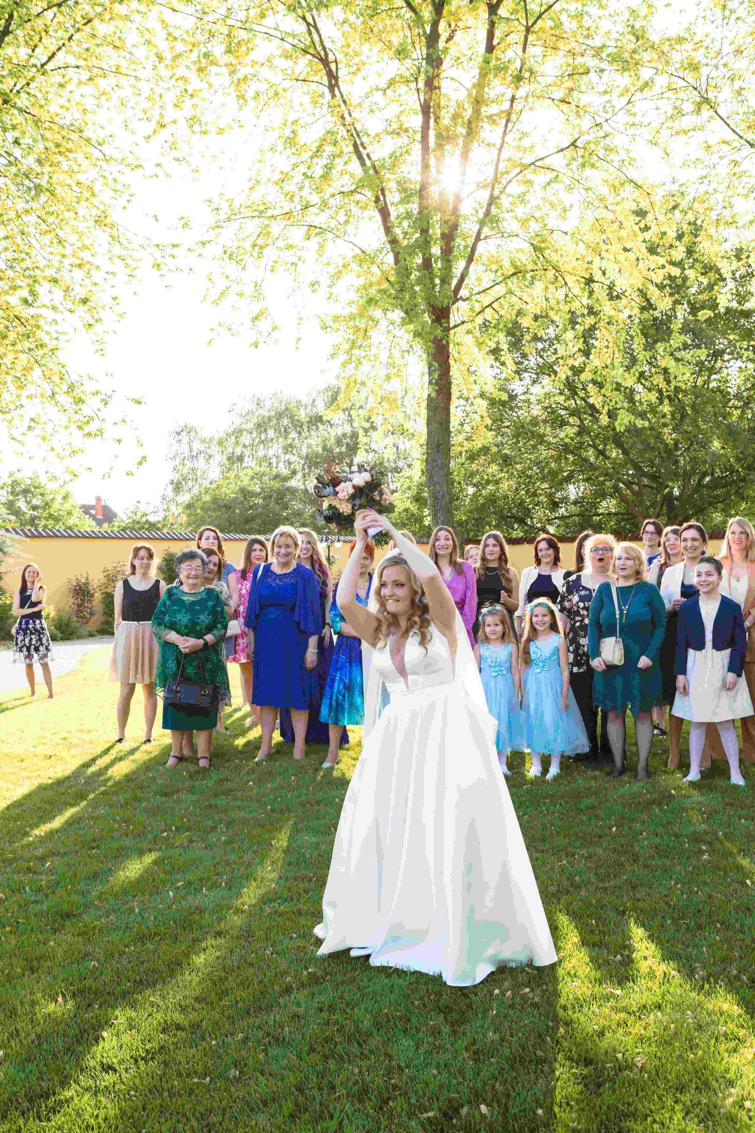 A group of wedding guests stand together on a grassy lawn during a sunny outdoor celebration.