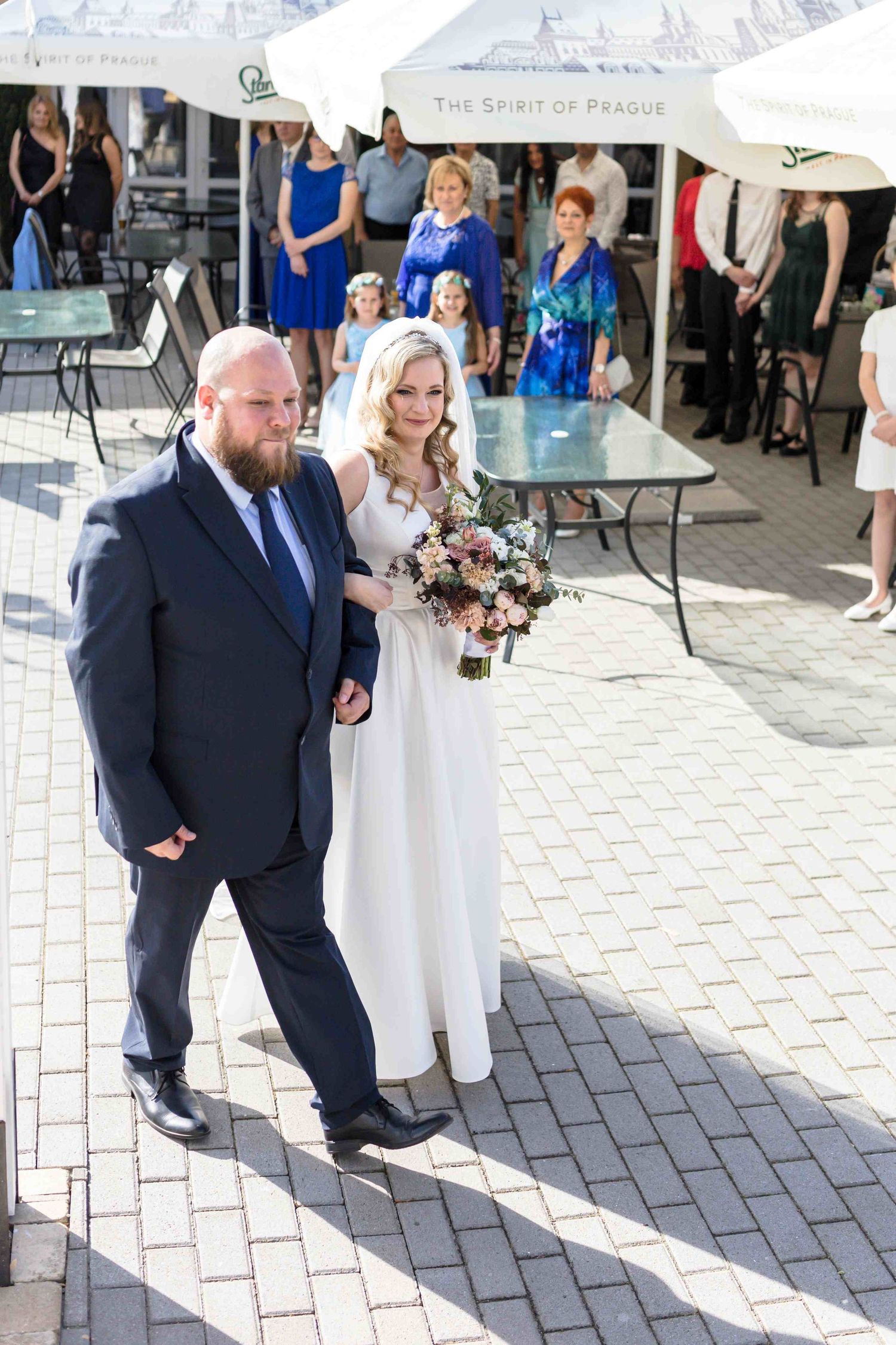 Wedding party members walk along a stone paved courtyard in formal attire during an outdoor celebration.
