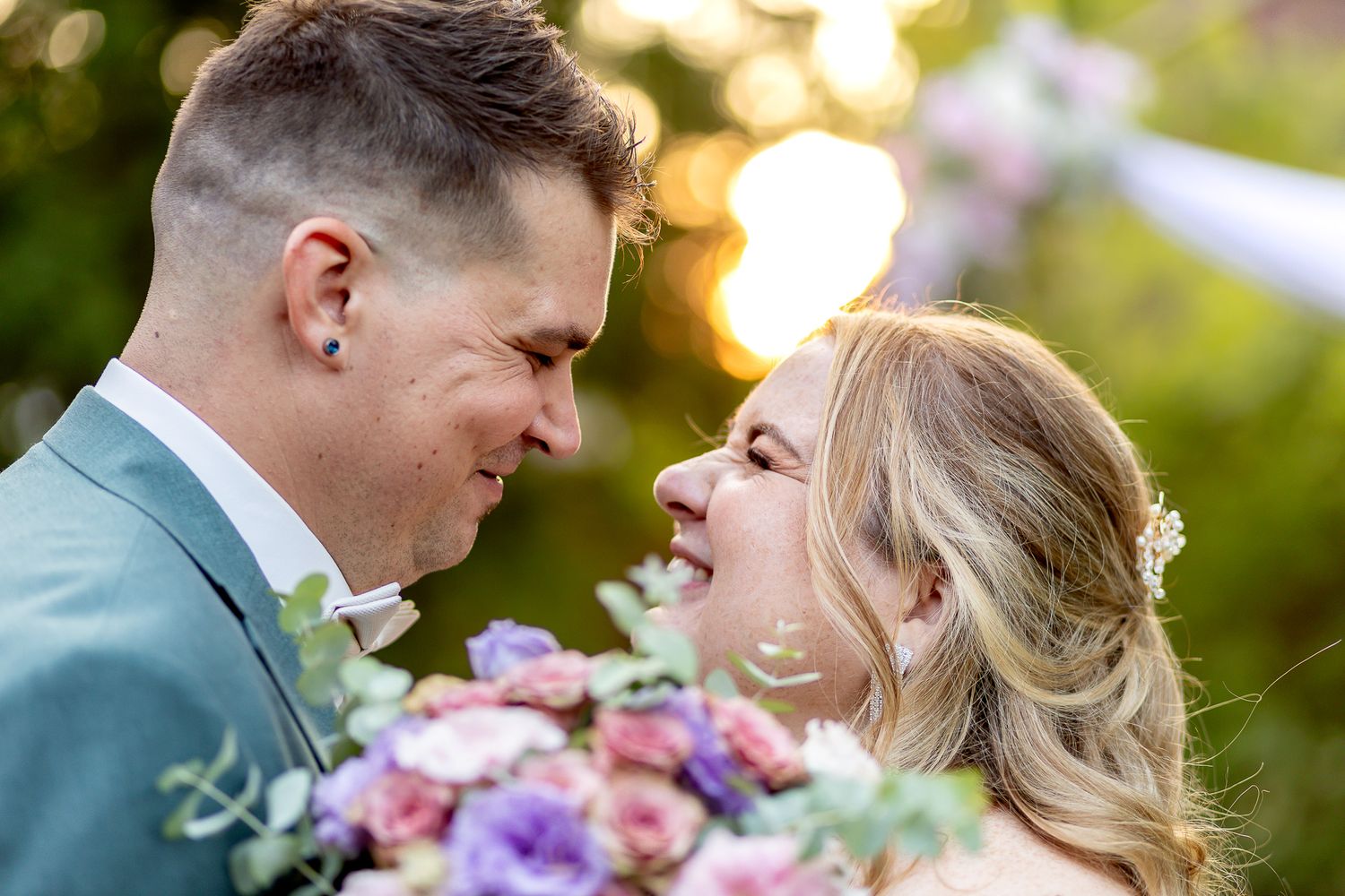 A romantic wedding moment captured in golden sunlight with a bouquet of purple and pink flowers between the couple.