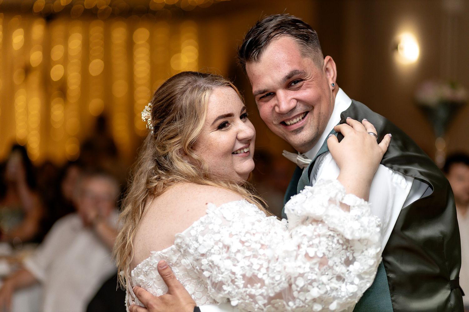 A couple shares a romantic dance at their wedding reception with warm lighting and guests in the background.