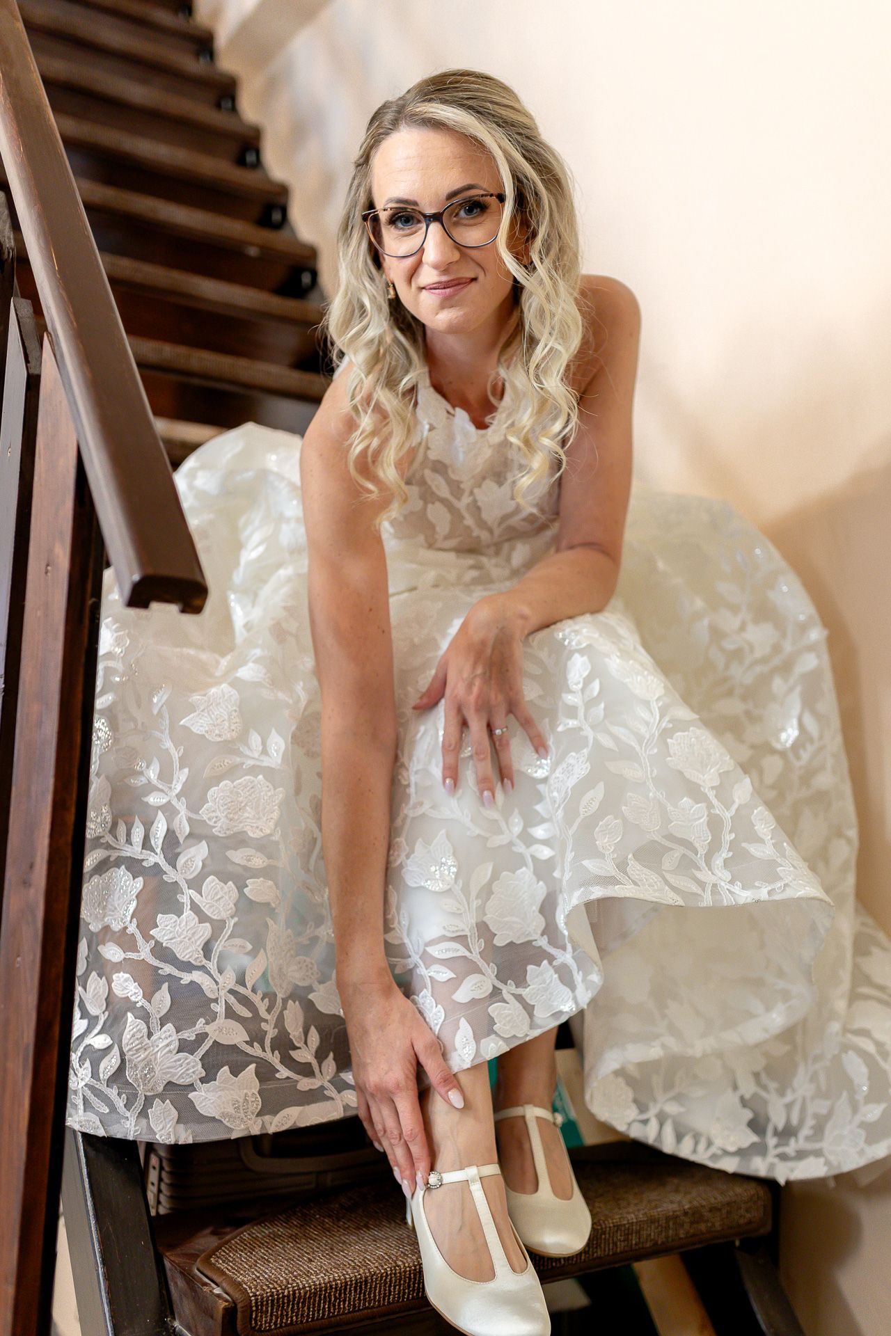 A bride in a white lace dress and glasses adjusts her shoe while sitting on wooden stairs.