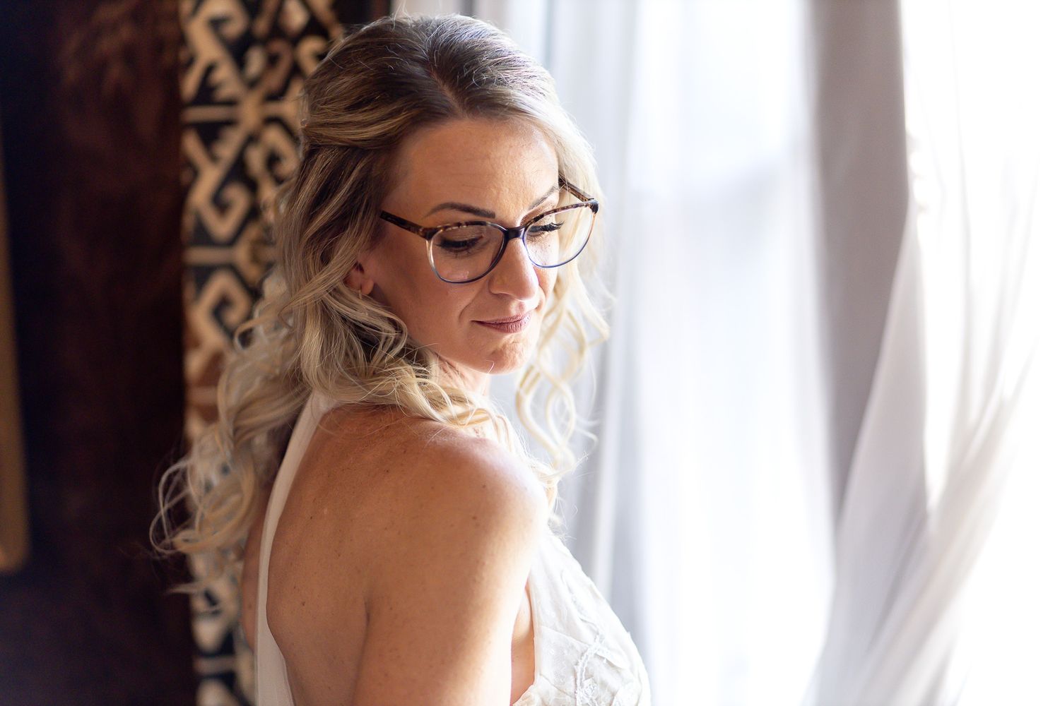 Bride wearing glasses and a white wedding dress looks out window with natural light streaming in.