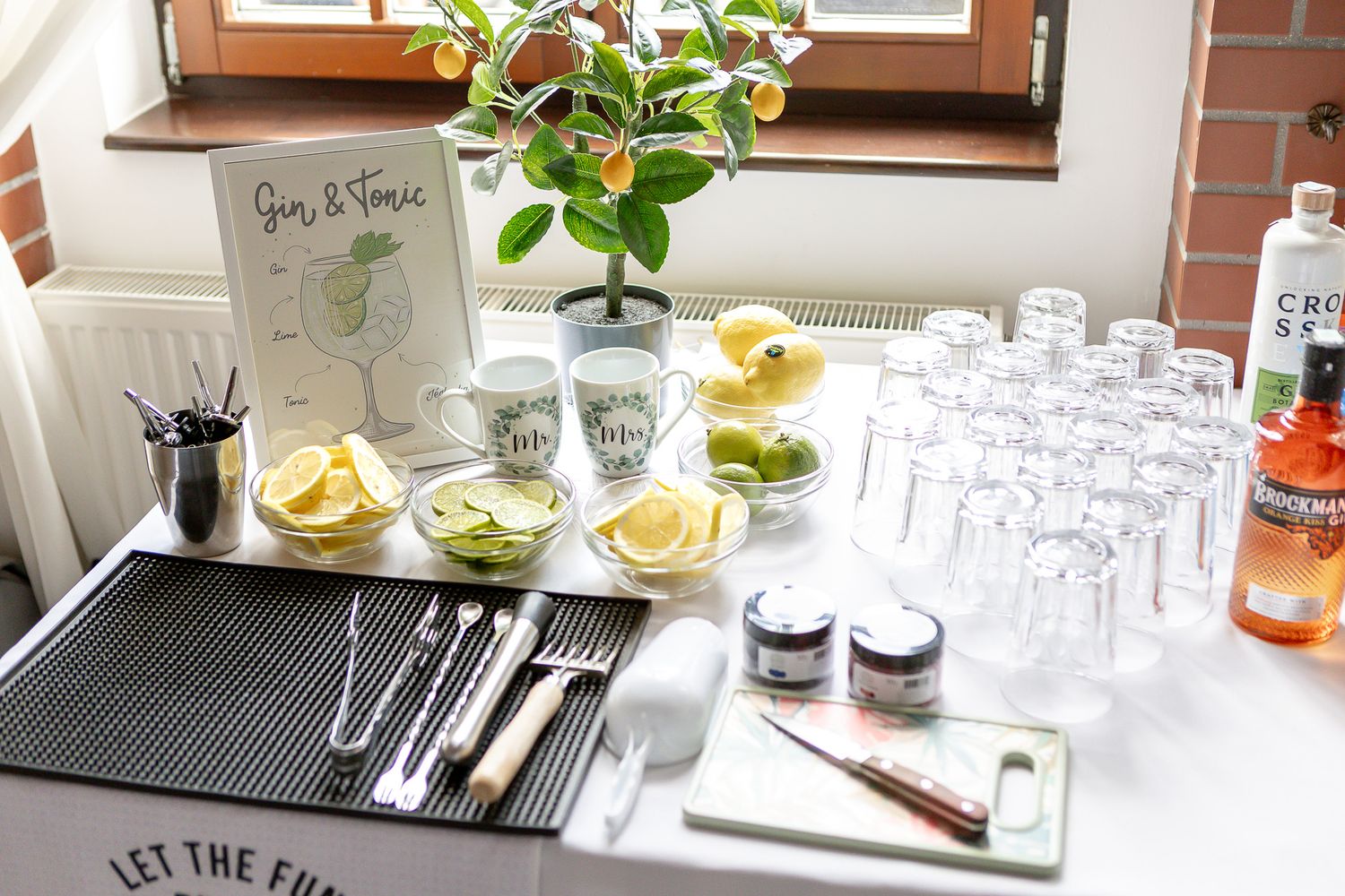 Various cocktail making supplies and glassware arranged on a bar counter with citrus garnishes and mixing tools.