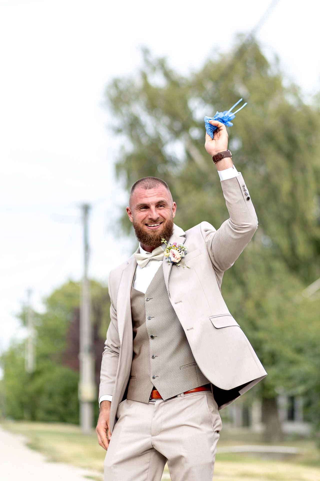 A person in a light beige suit raises a blue butterfly in celebration at an outdoor event.