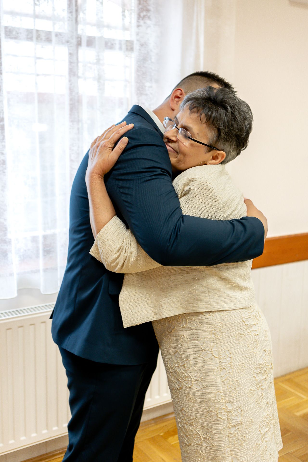 Two people share an emotional embrace in a bright room with white curtains and wooden flooring.