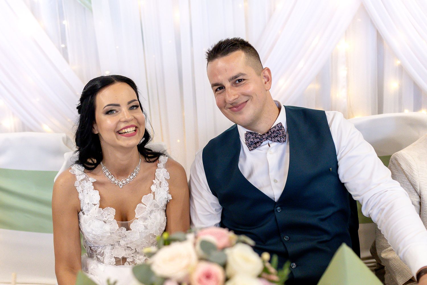 A couple in formal wedding attire share a romantic moment at their reception table with white decor and floral arrangements.