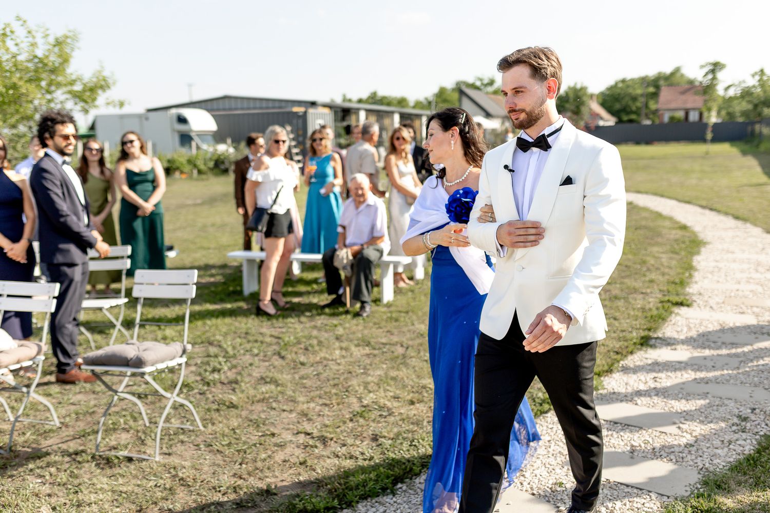 Wedding guests gather on pathway at outdoor venue with white chairs set up on lawn.