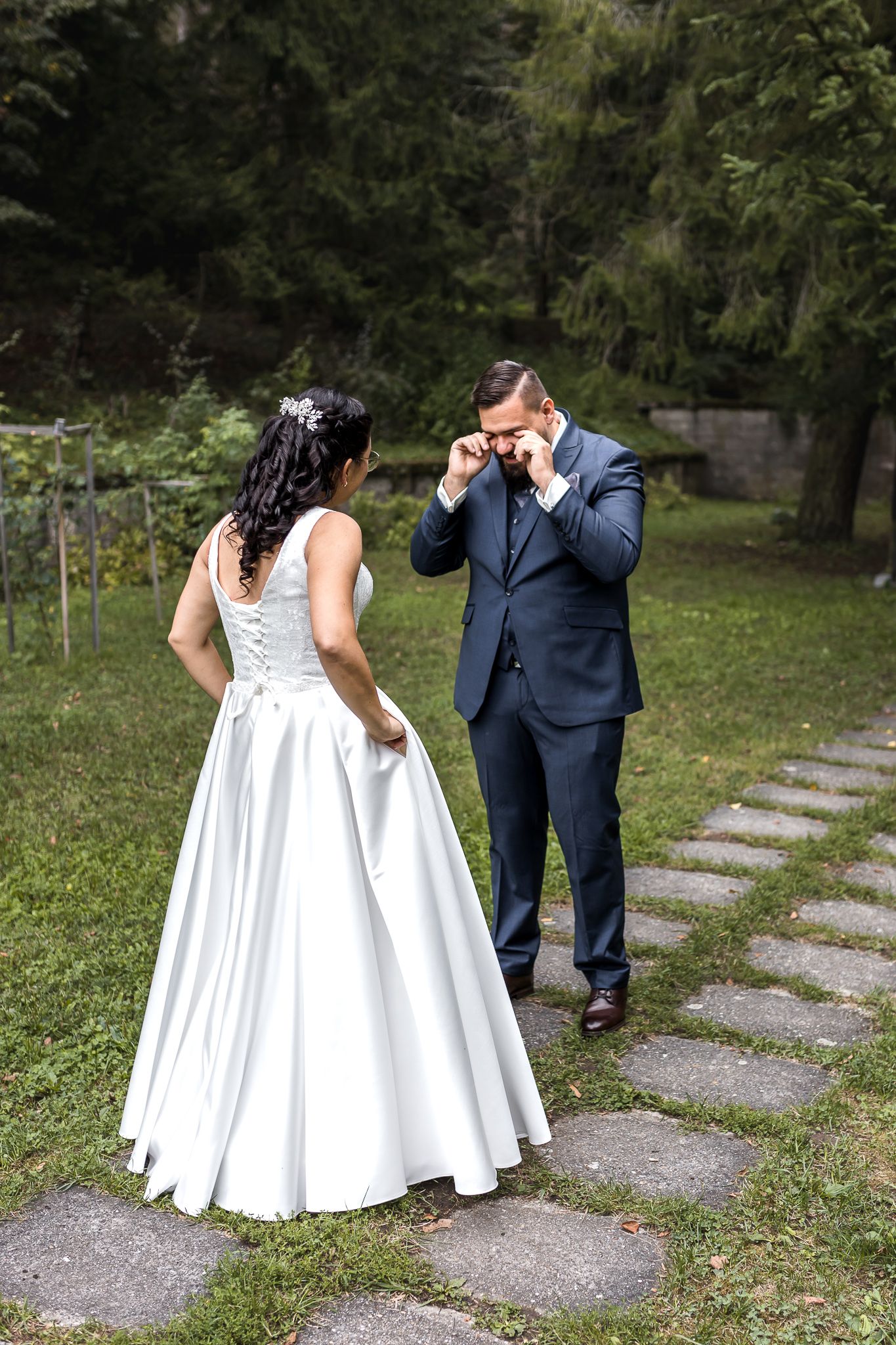 A couple in wedding attire walks along a stone path in a garden setting.