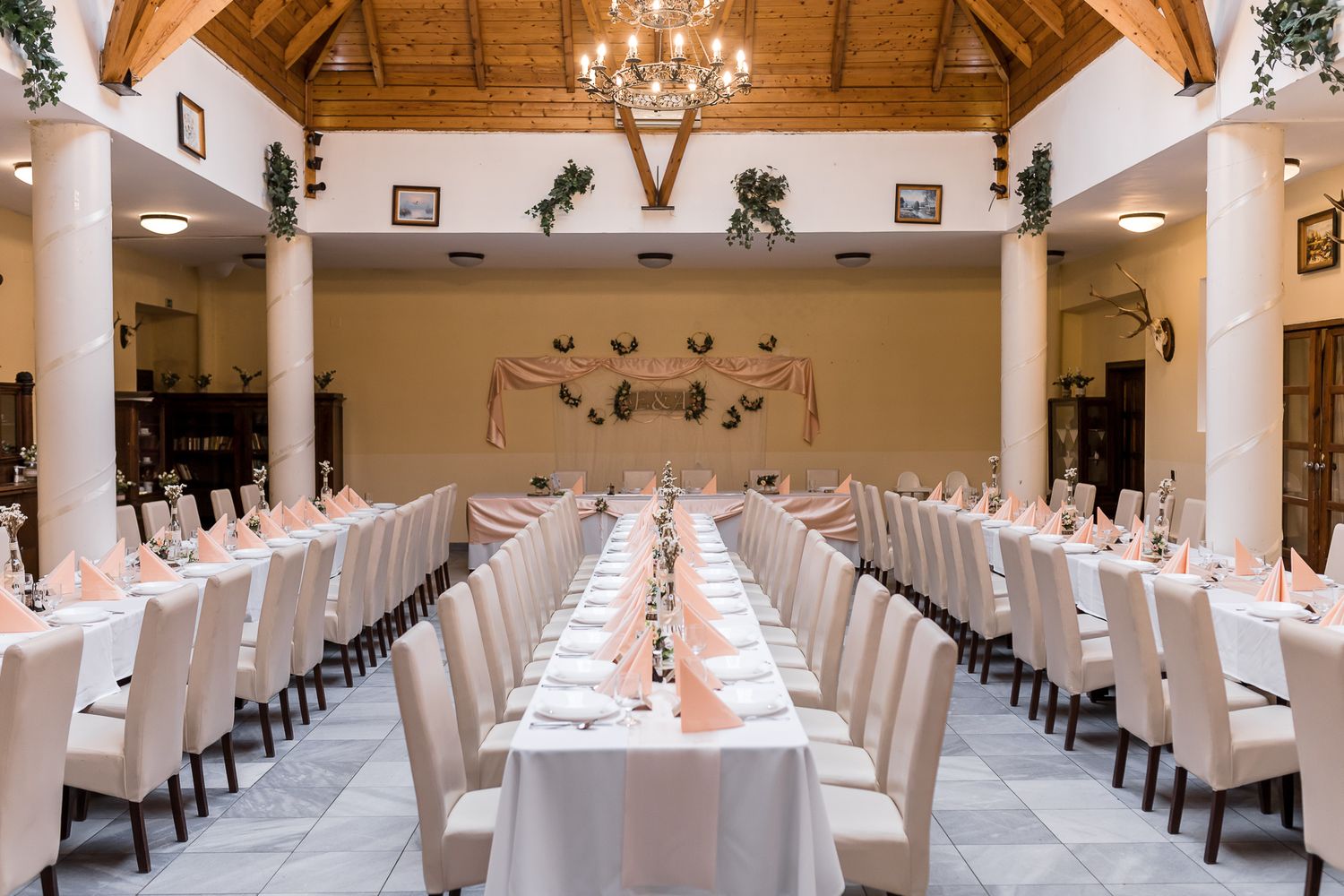An elegant wedding reception venue with long white tables, chandeliers, and vaulted wooden beam ceilings.