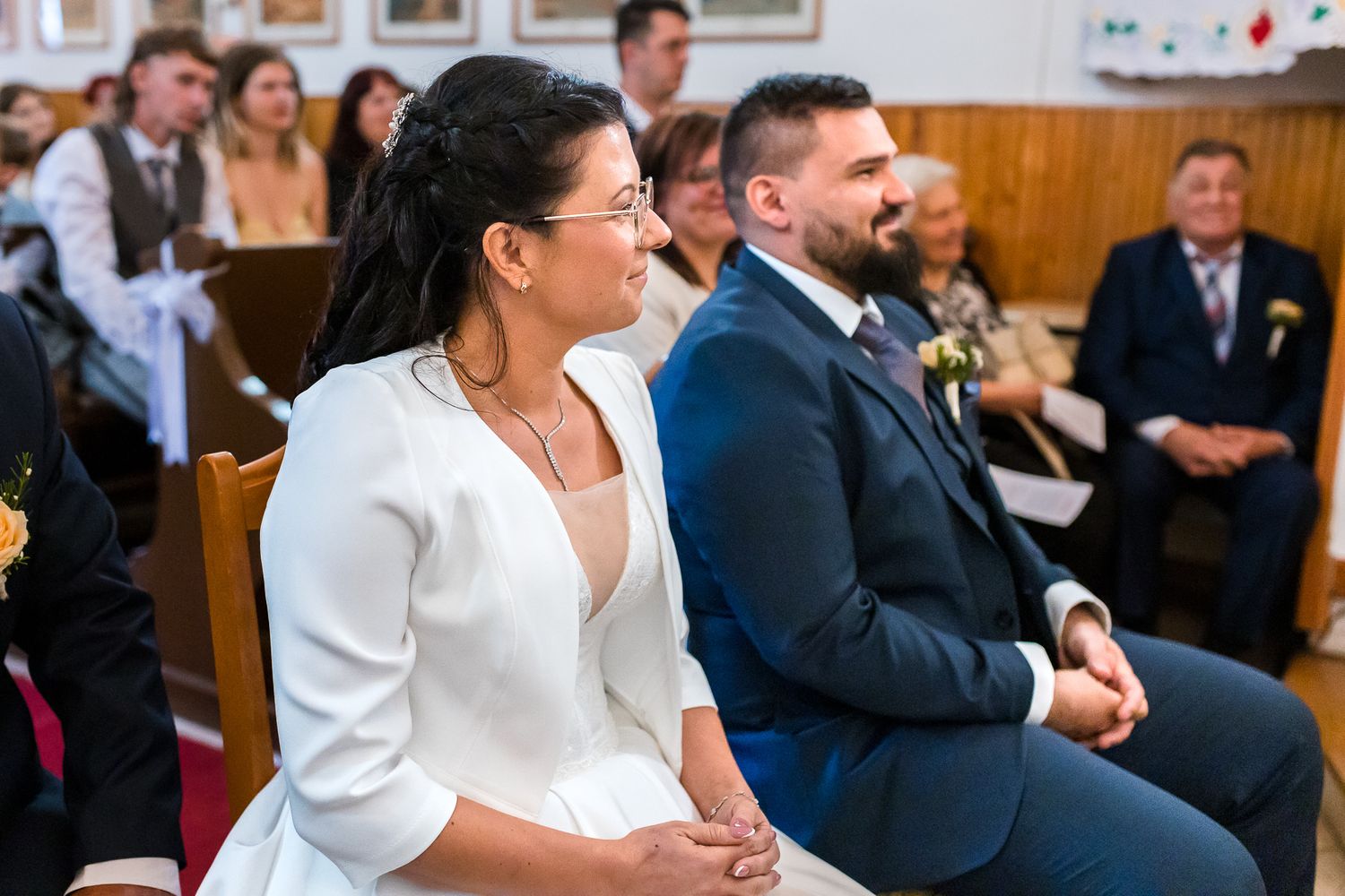 A couple laughs joyfully during their wedding ceremony while seated in wooden church pews.