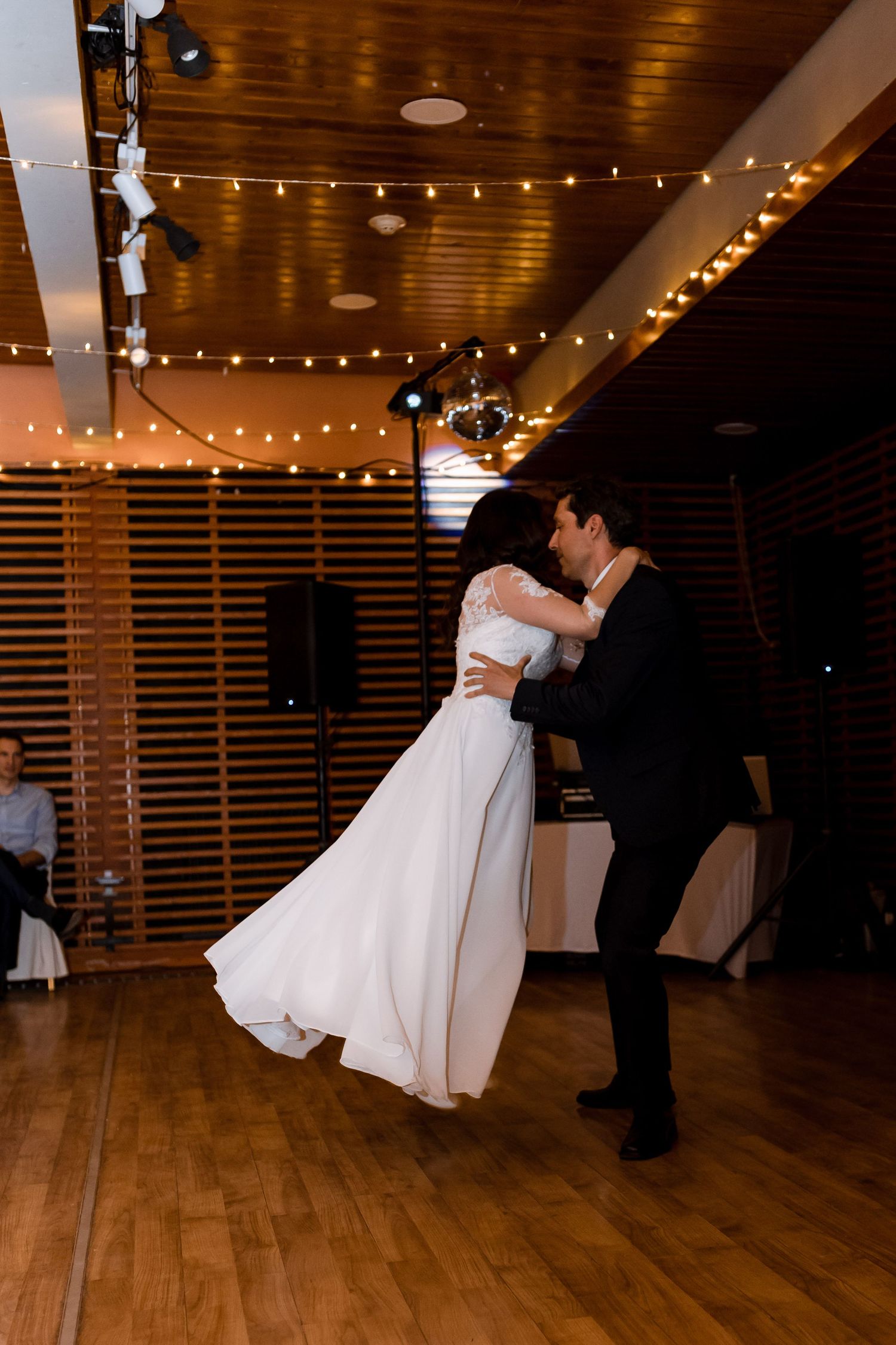 A couple performs a romantic dip dance move on a wooden dance floor under warm string lights at a wedding reception.