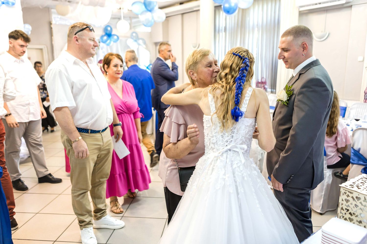 Wedding celebration scene with guests gathered around a couple in formal attire at a venue decorated with blue balloons.