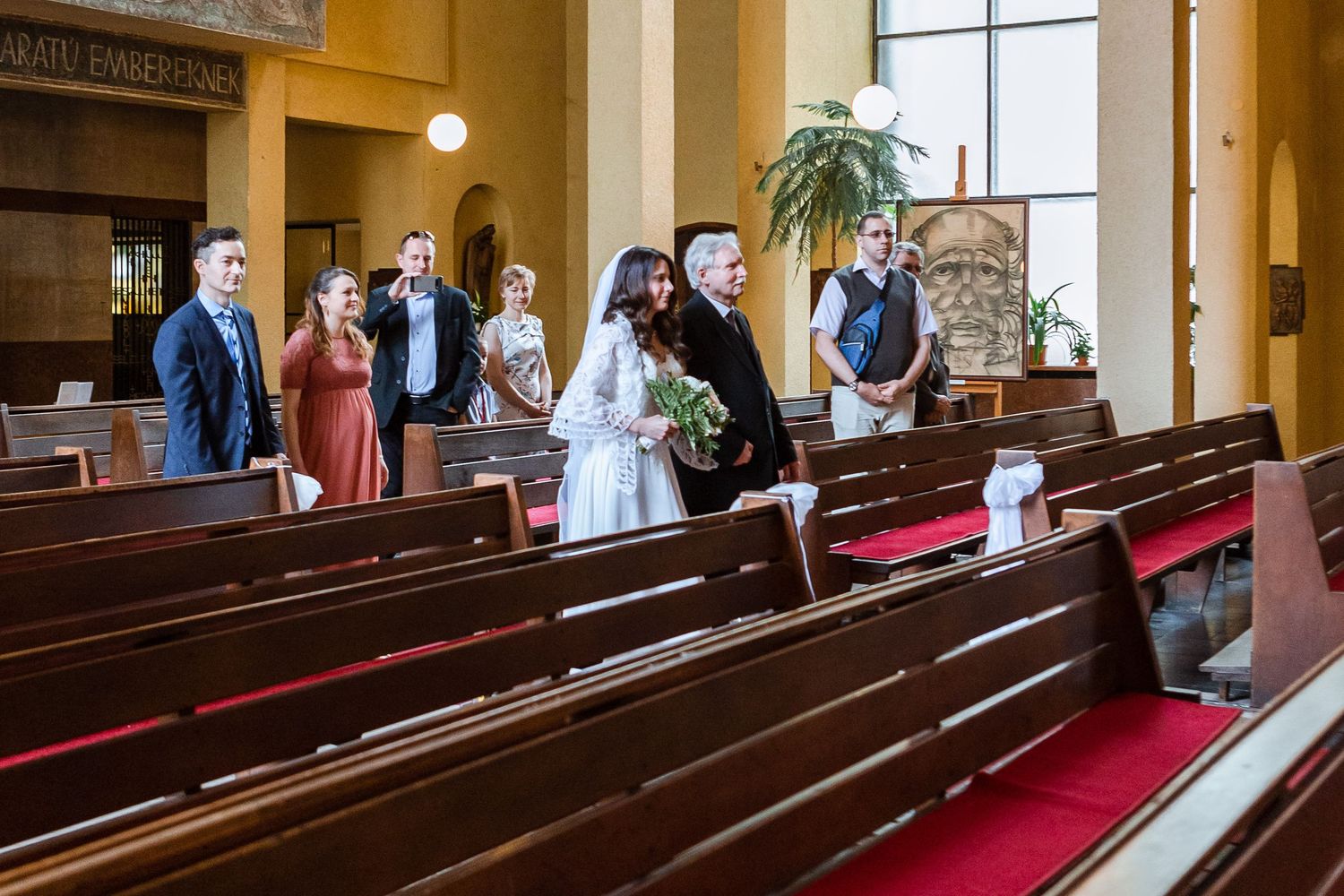 People gather in wooden church pews with red cushions while light streams through tall windows in a chapel interior.
