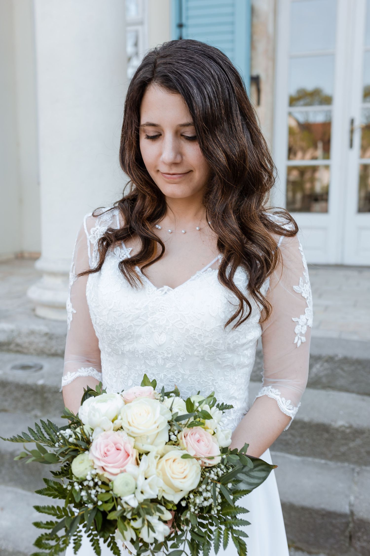 A bride in a white lace wedding dress holds a beautiful bouquet of pink and white roses with greenery.