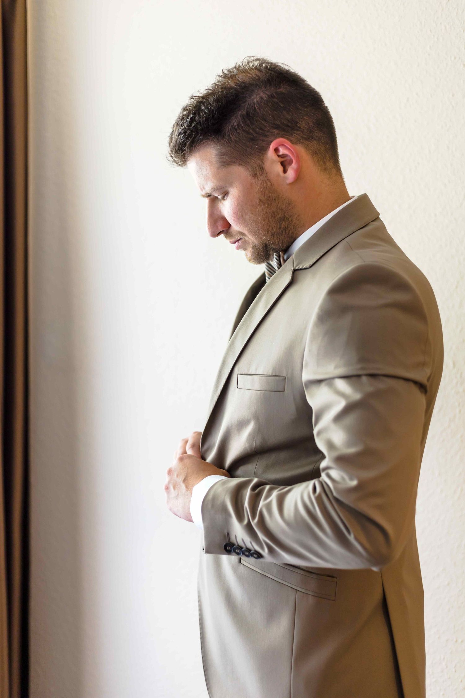 A person in a light beige suit adjusts their jacket while standing against a white wall.