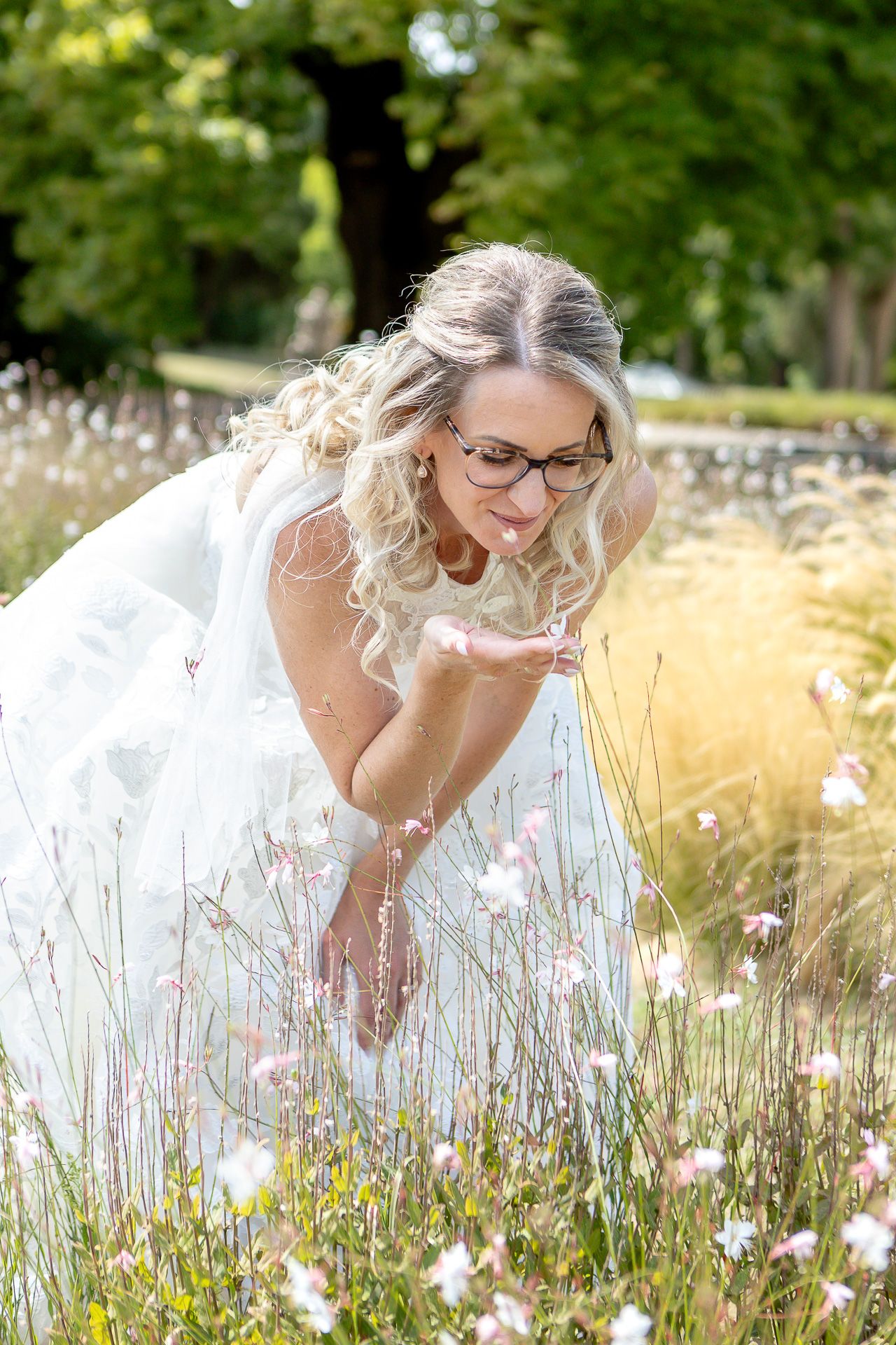 A person in glasses and white dress poses gracefully in a summer meadow with tall grass and wildflowers.