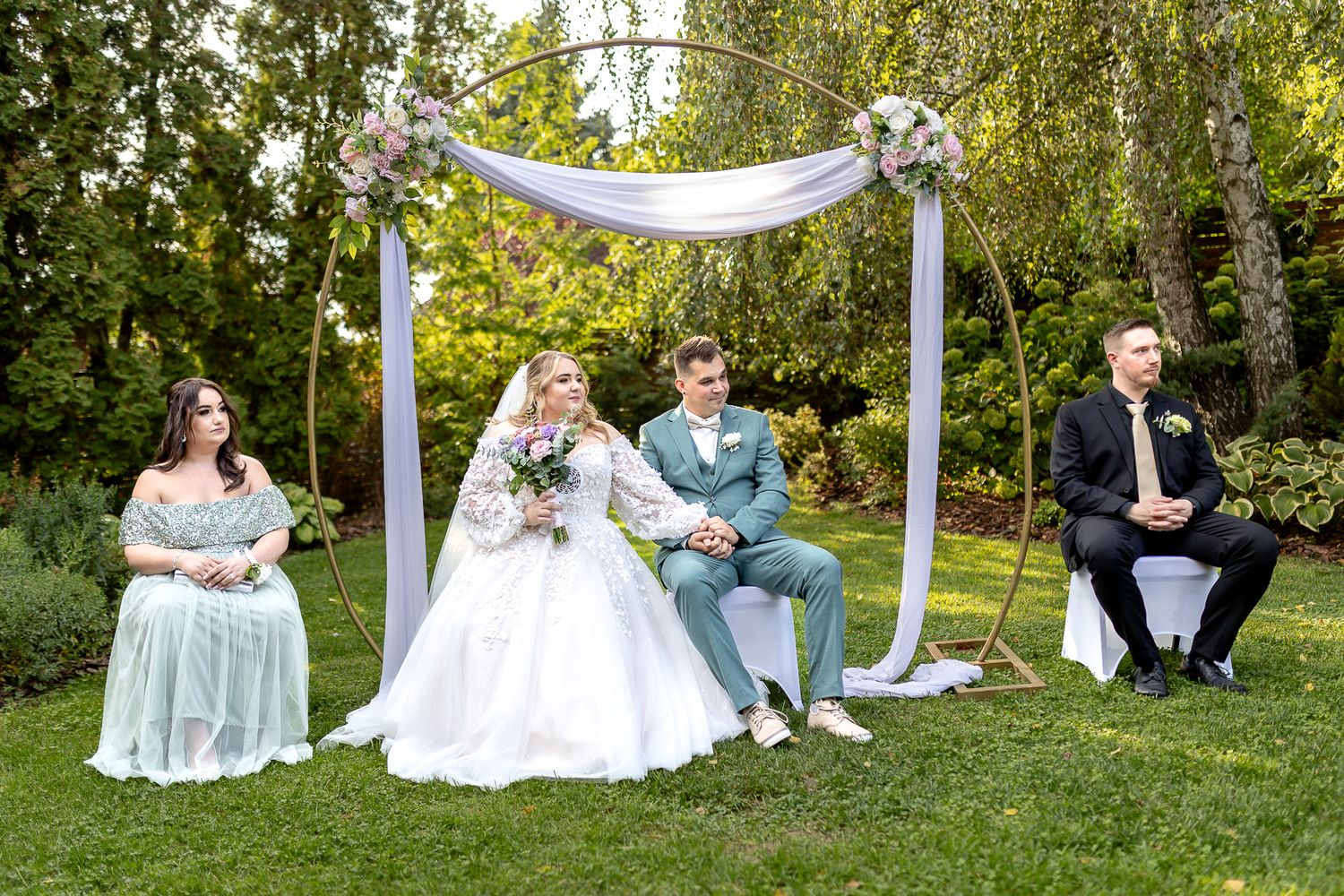 A small outdoor wedding ceremony setup with a white draped arch and guests seated on chairs in a garden setting.