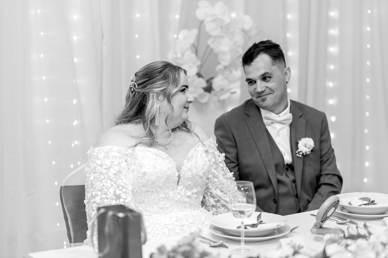 Wedding guests seated at a decorated reception table with white linens and twinkle lights in the background.