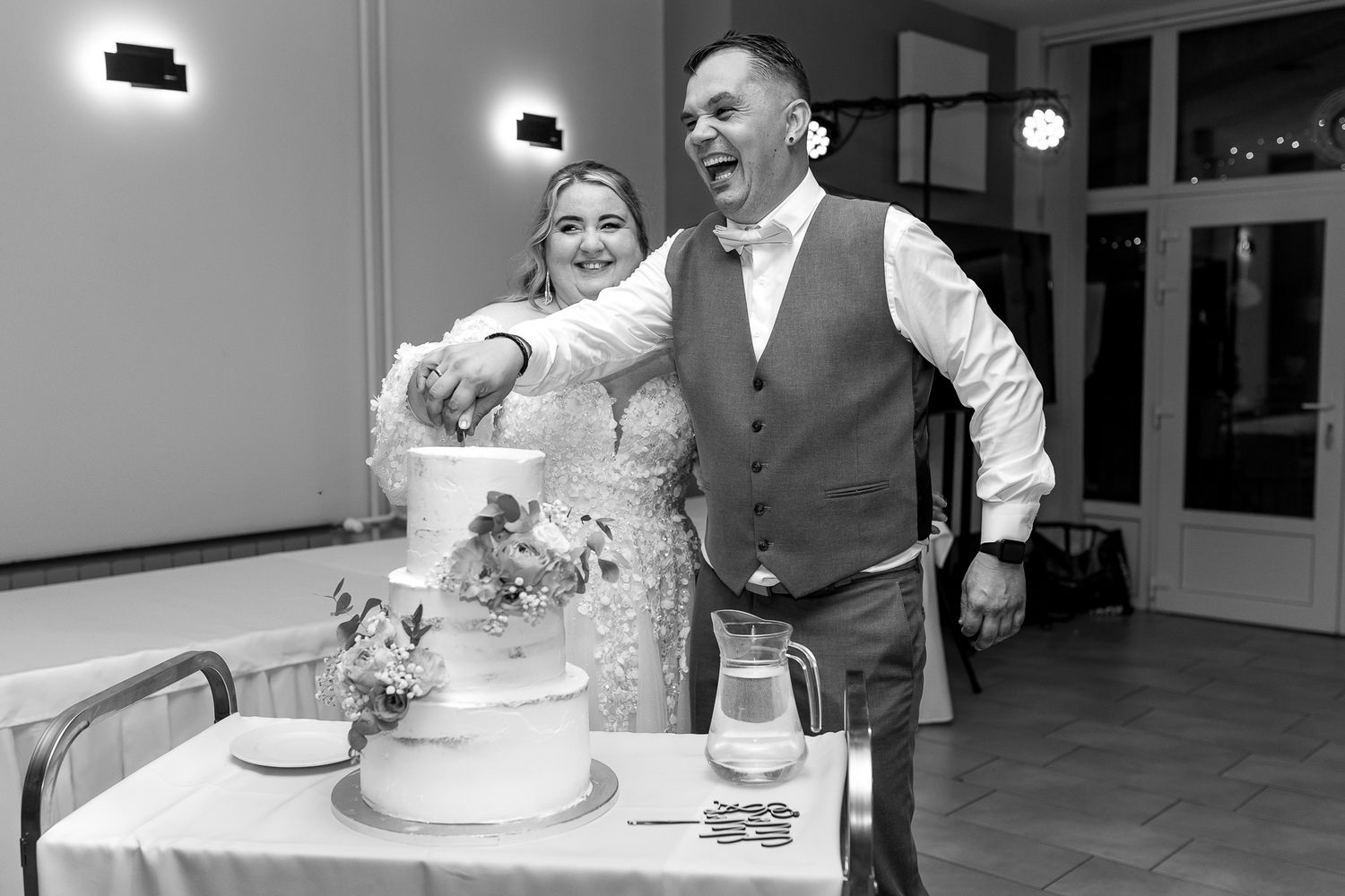 Black and white photo of a wedding couple cutting their tiered white cake at the reception.