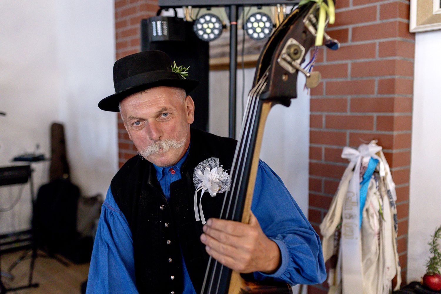 Musician in black hat and blue shirt plays upright bass against brick wall backdrop during performance.