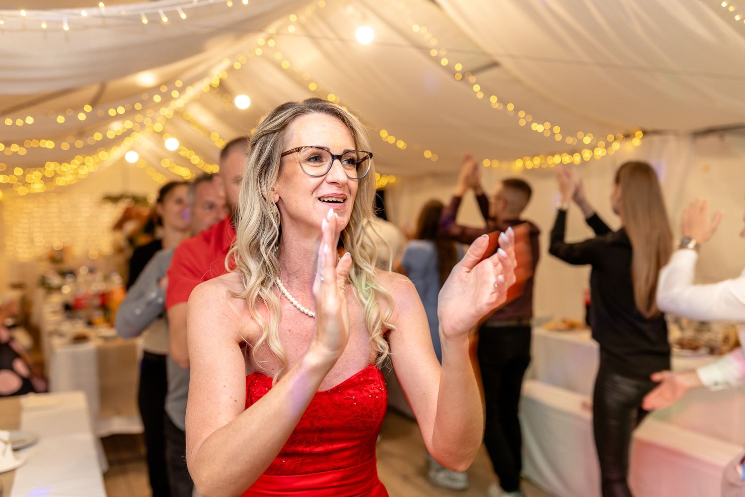 A group of people dancing and celebrating at an indoor event decorated with string lights and white fabric.