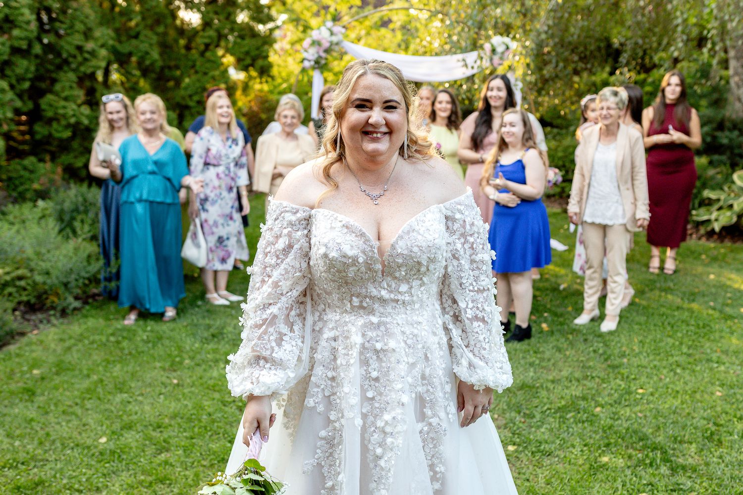 A wedding celebration on a sunny day with attendees gathered on a green lawn wearing colorful formal attire.