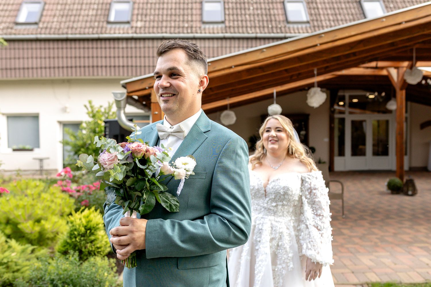 A couple in wedding attire share a joyful moment outside a building with a tiled roof and beautiful garden.