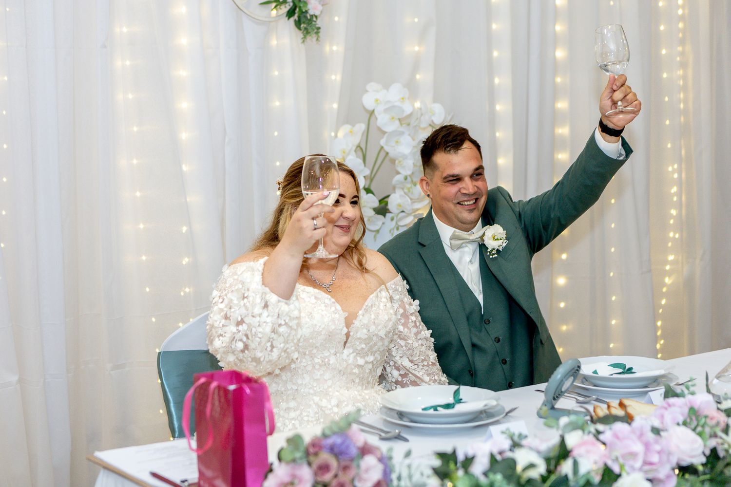 A wedding couple and guests celebrate during a reception dinner with twinkle lights glowing in the background.