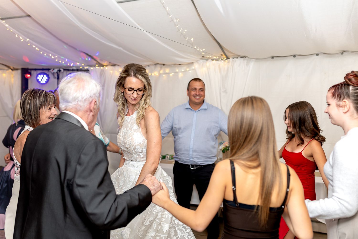 Wedding reception guests dancing and socializing under white tent with string lights and decorative lighting.