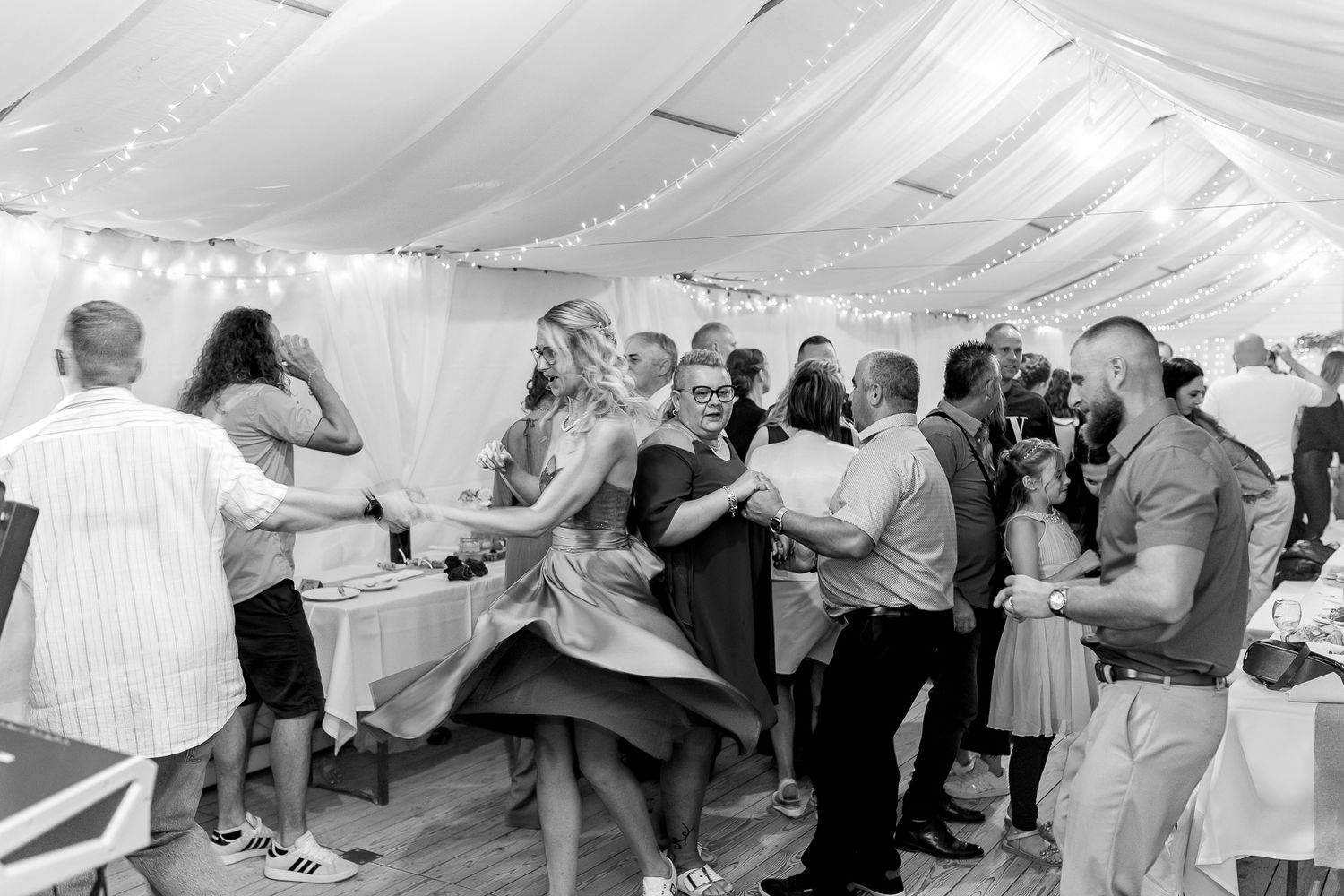 Wedding guests dancing together in a white tent decorated with fabric draping and string lights.