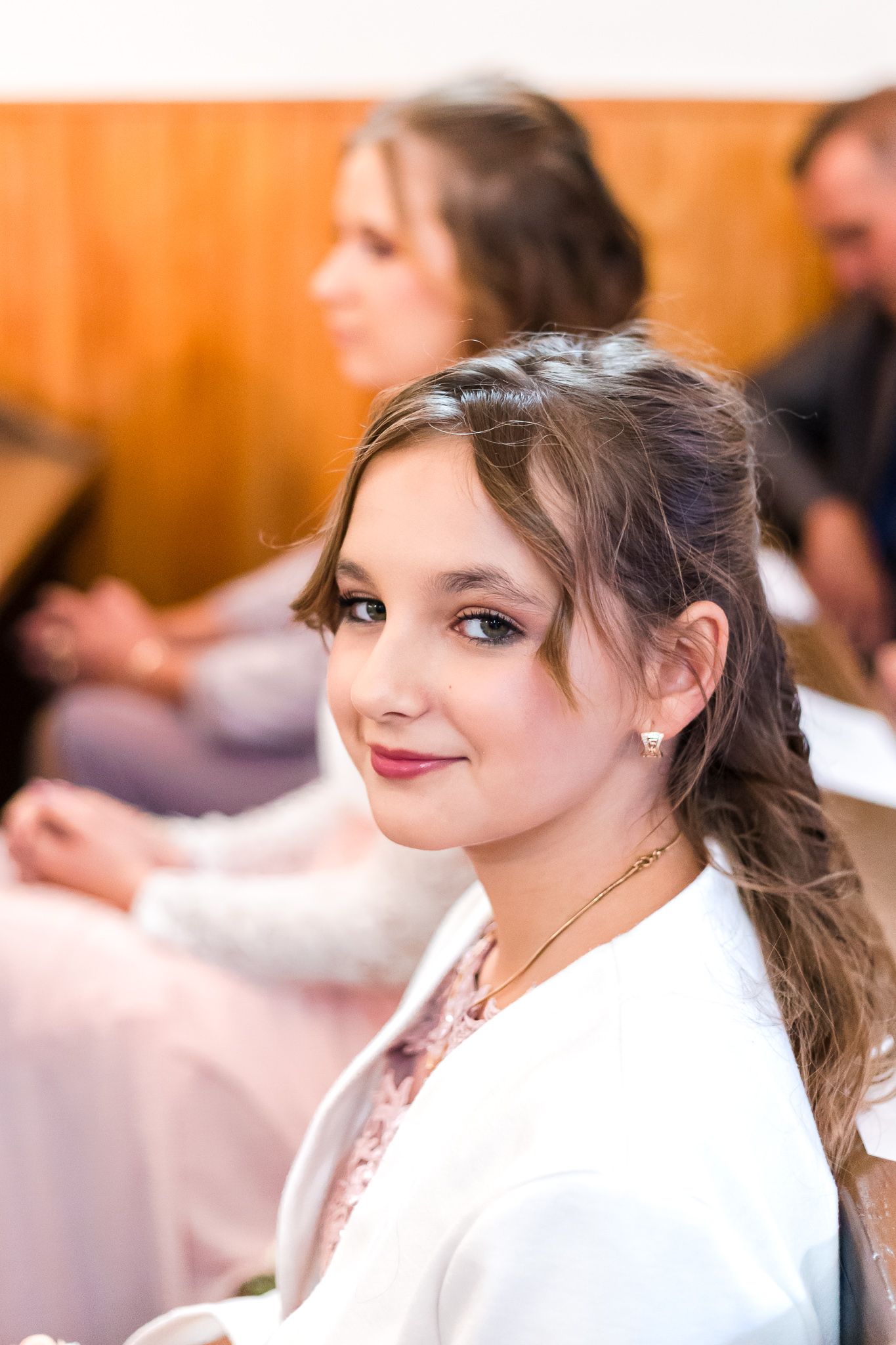 A person in white attire smiles warmly while attending an indoor event with soft lighting and blurred background.