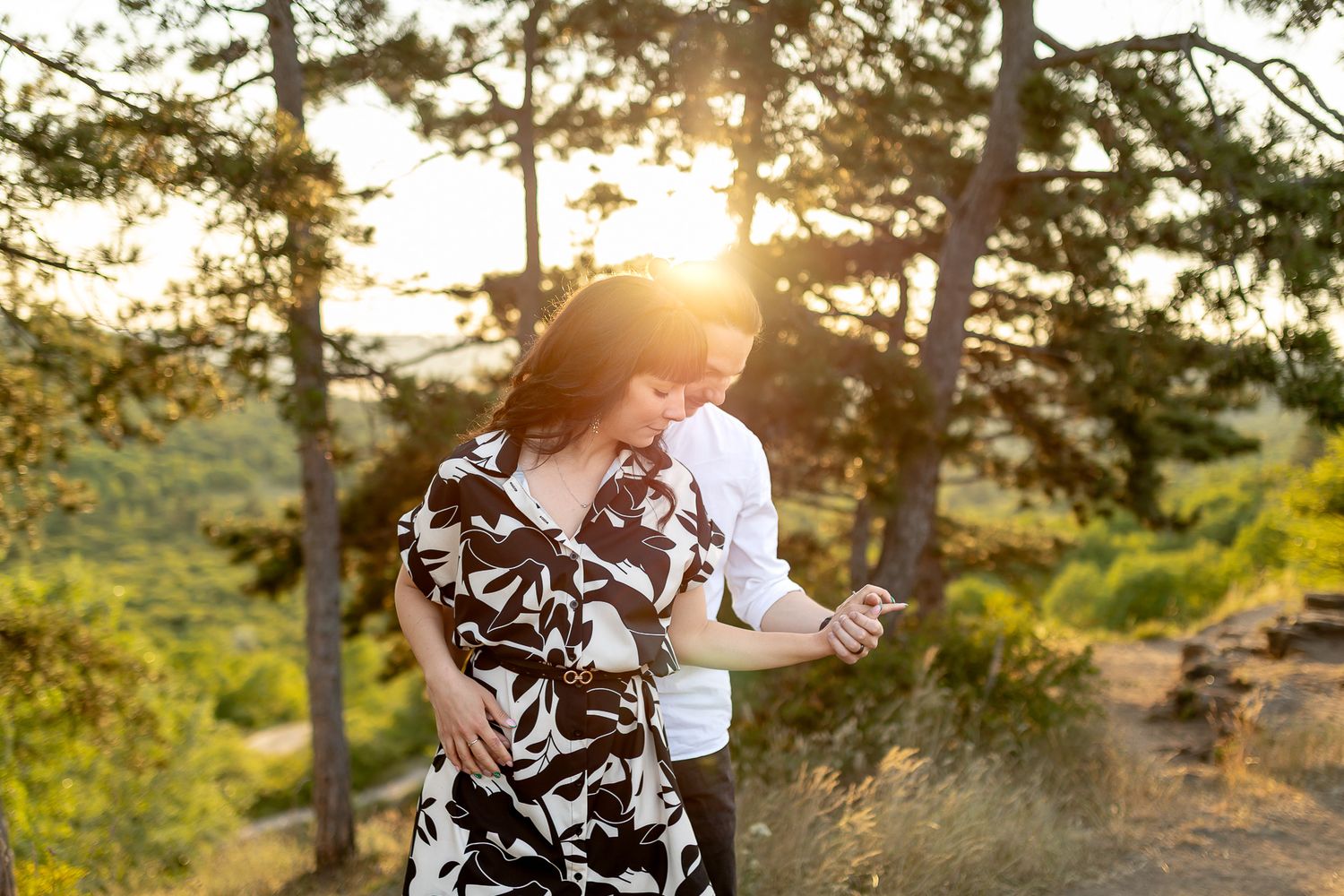 A couple shares a tender moment outdoors with sunlight filtering through trees in the background.