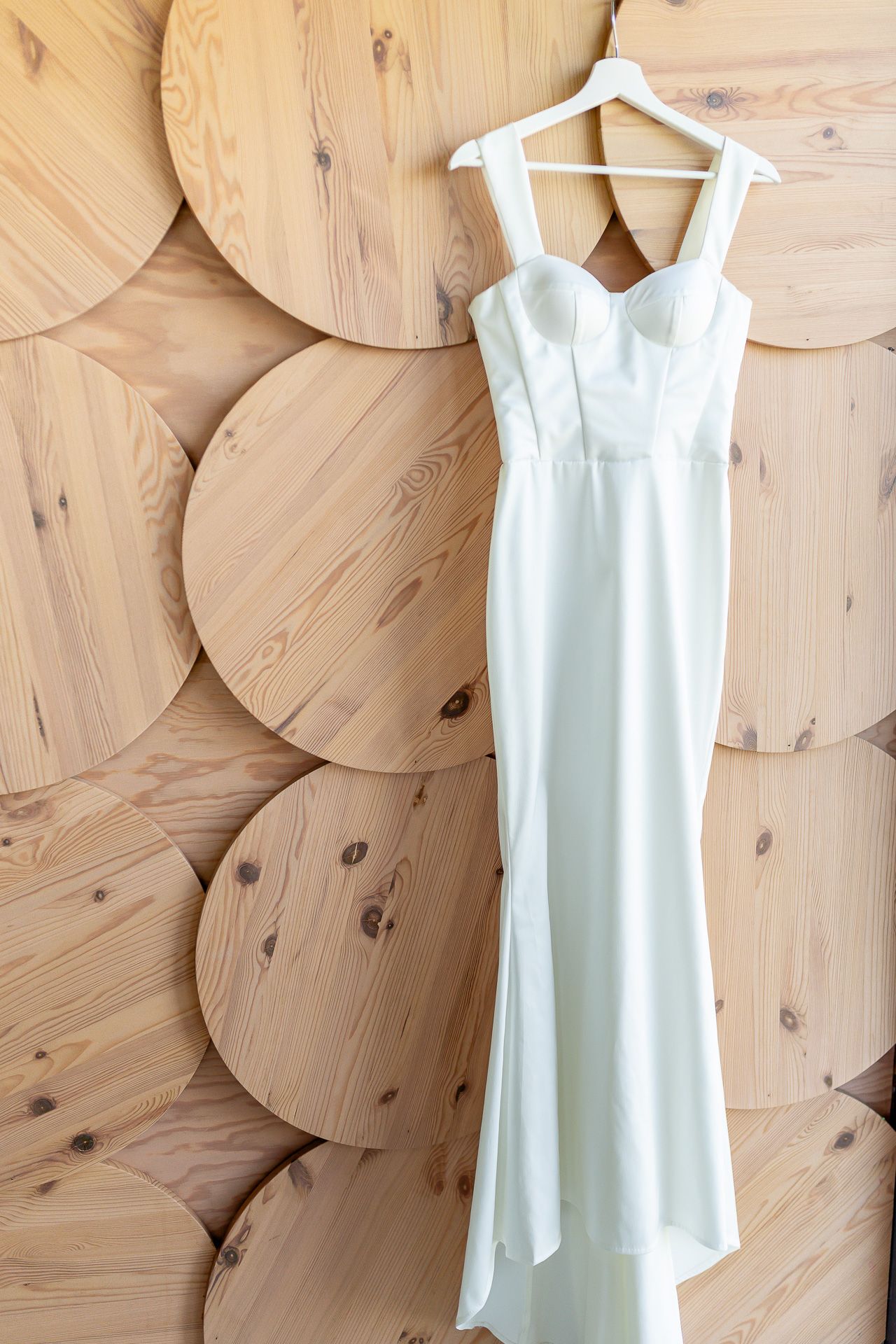 An elegant white wedding dress with straps hangs against a wooden circular wall pattern.