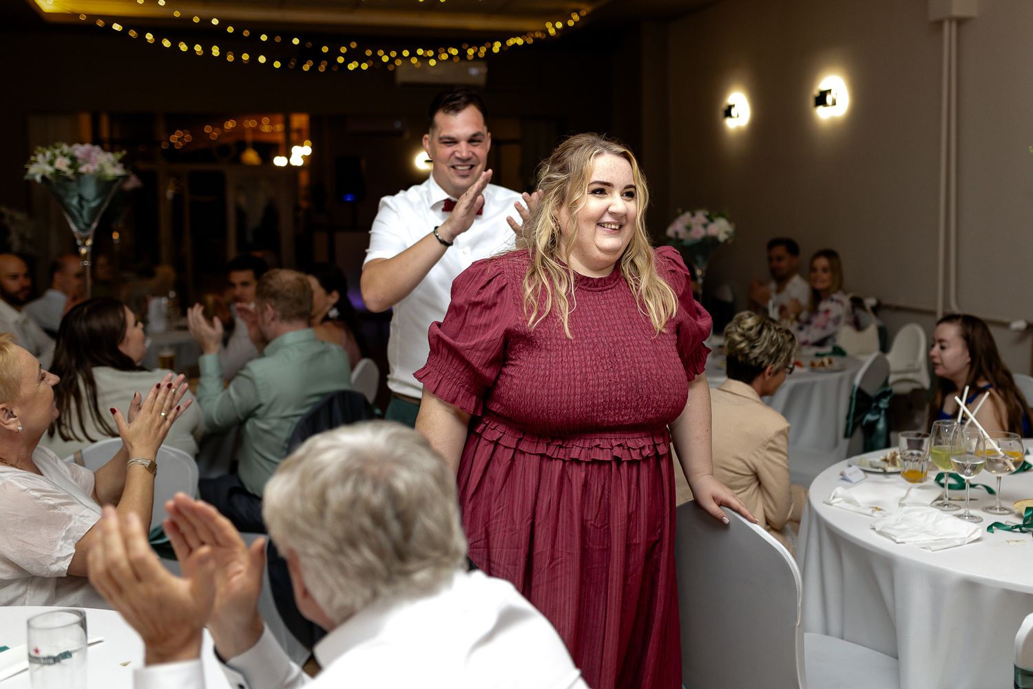 Wedding reception guests dance and celebrate together under decorative string lights at an indoor venue.