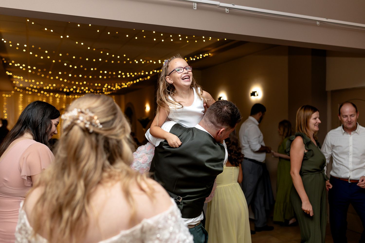 Wedding guests dance and celebrate on a dimly lit dance floor under twinkling string lights at an evening reception.
