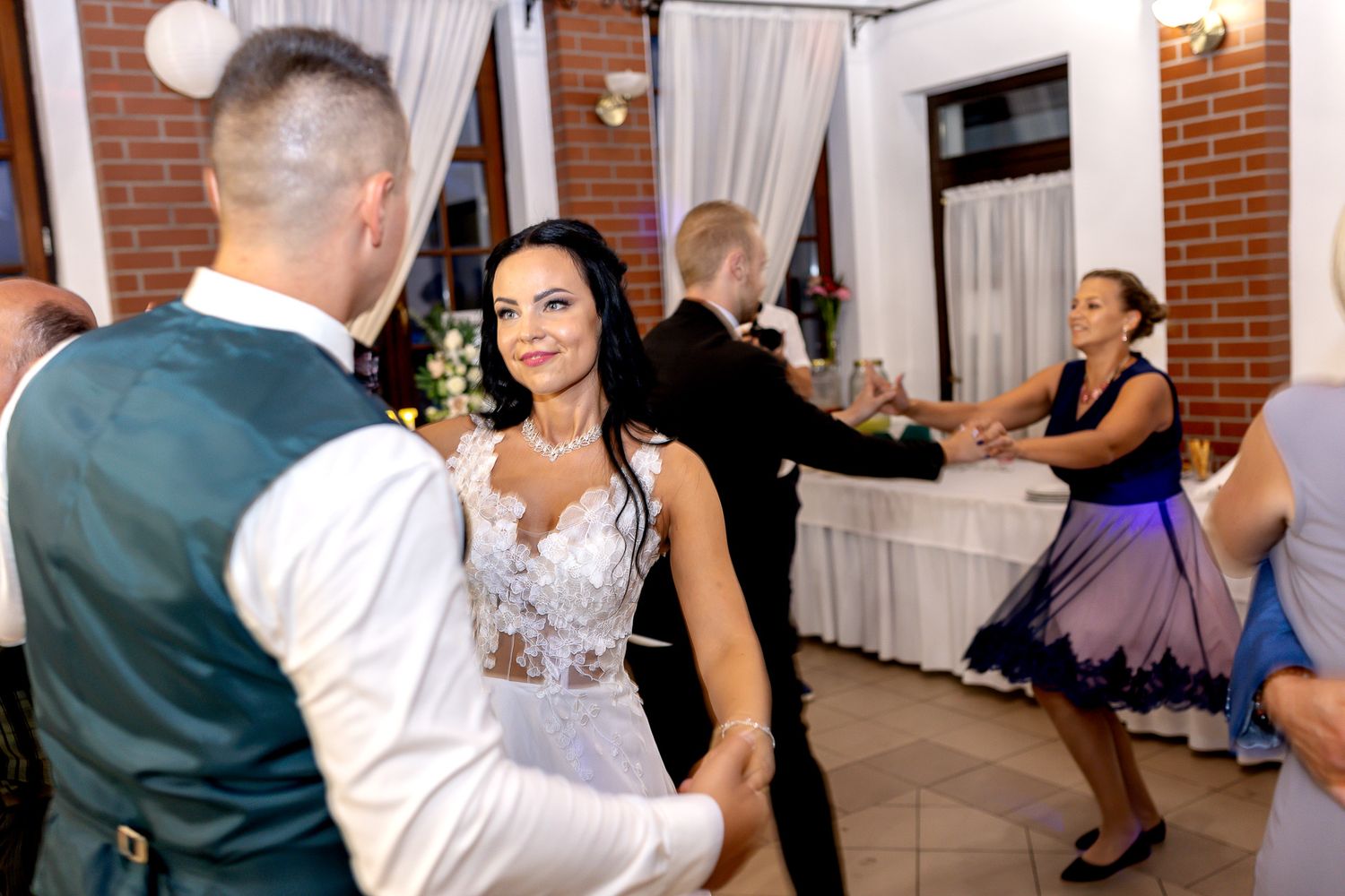 Wedding guests dance and celebrate together on the dance floor at an indoor reception venue with brick walls.