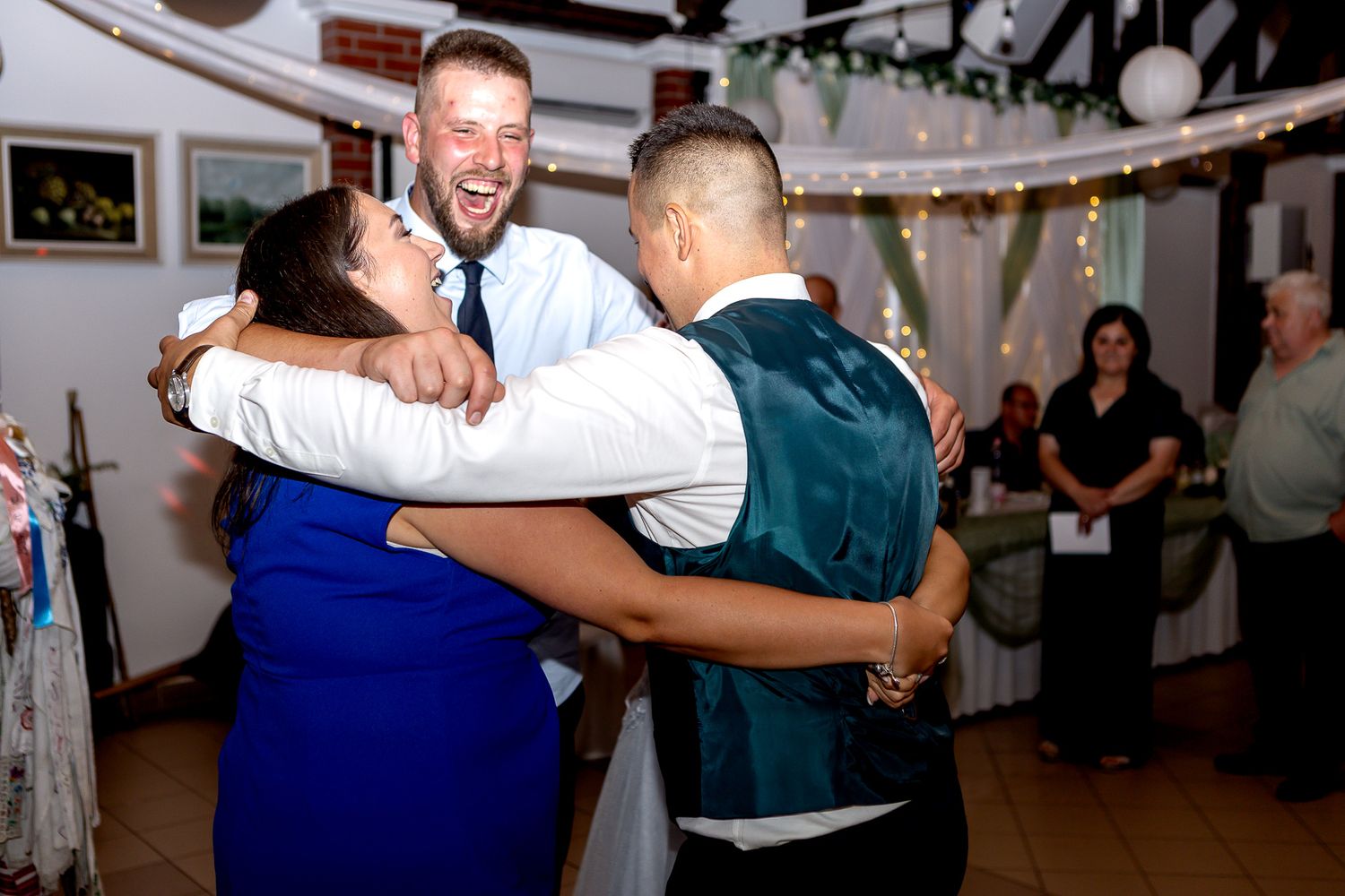 Wedding guests dance and embrace on a festive dance floor decorated with string lights during a reception celebration.