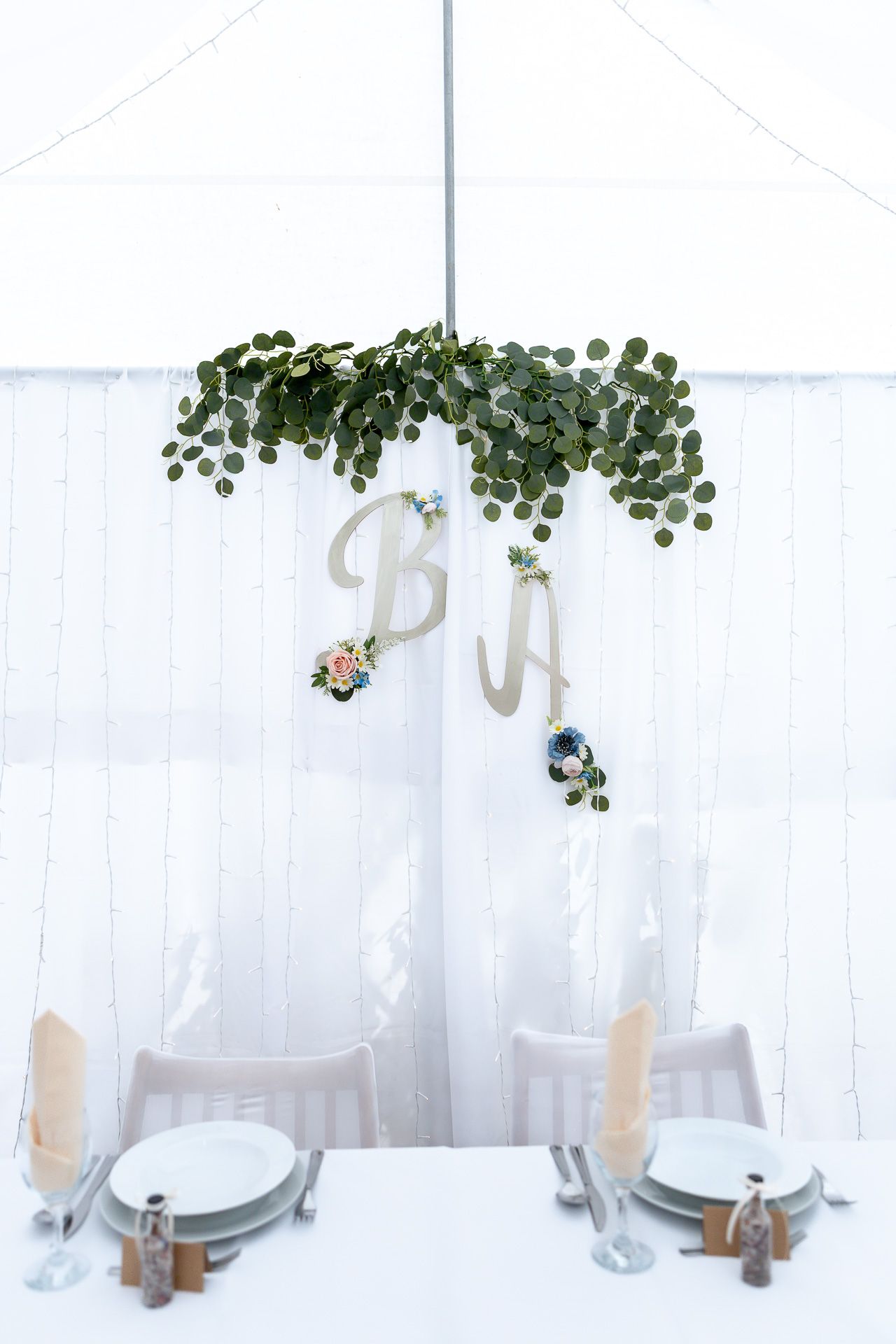 White plates and wine glasses set on table with delicate green vine decorations and sheer curtain backdrop.