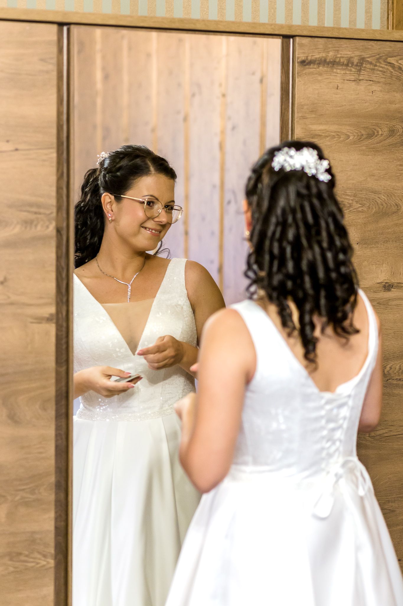 Two people in white dresses stand together in a room with wooden walls during what appears to be a wedding preparation.