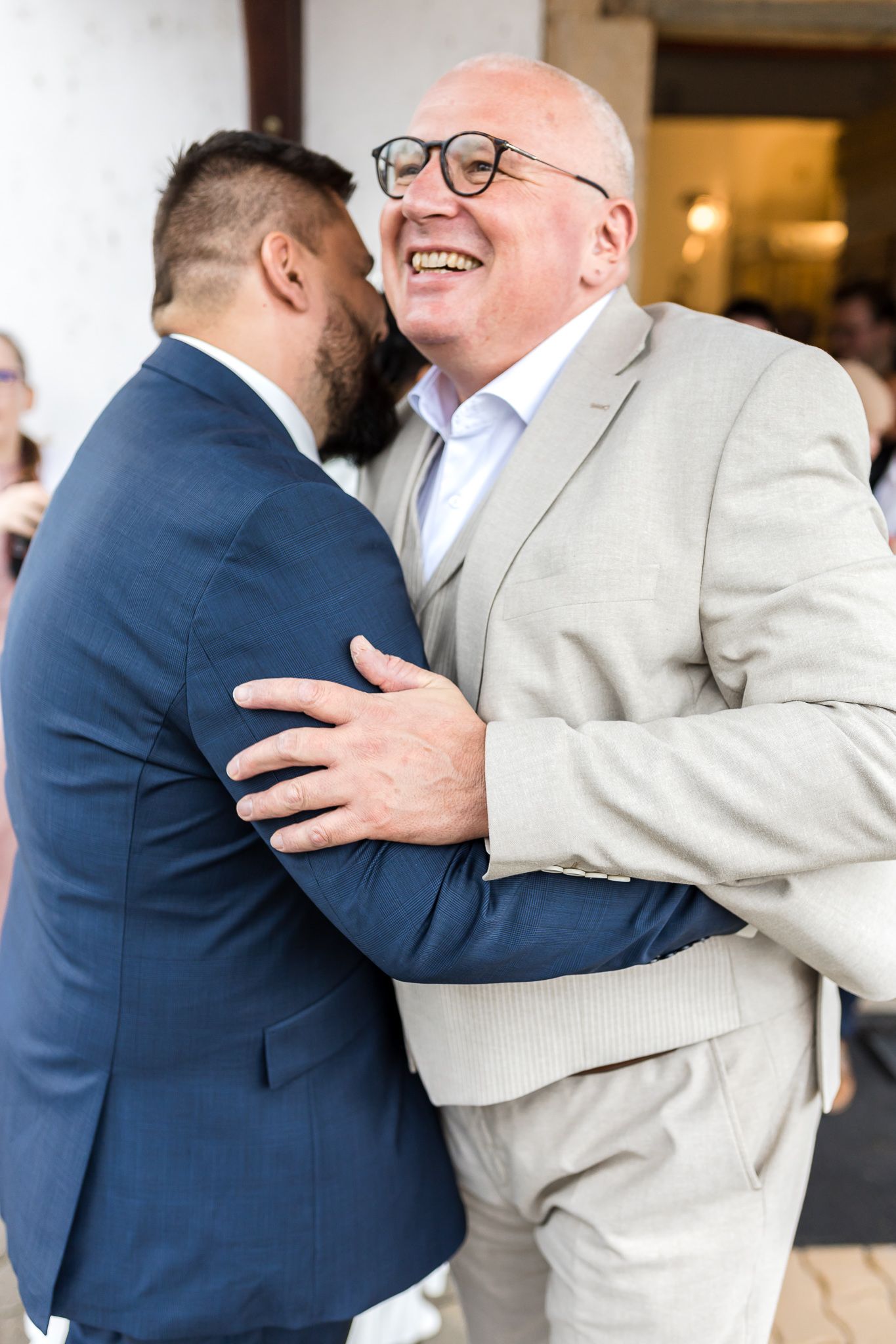 Two men share an emotional embrace at a formal event, one wearing a navy suit and one in a beige jacket.