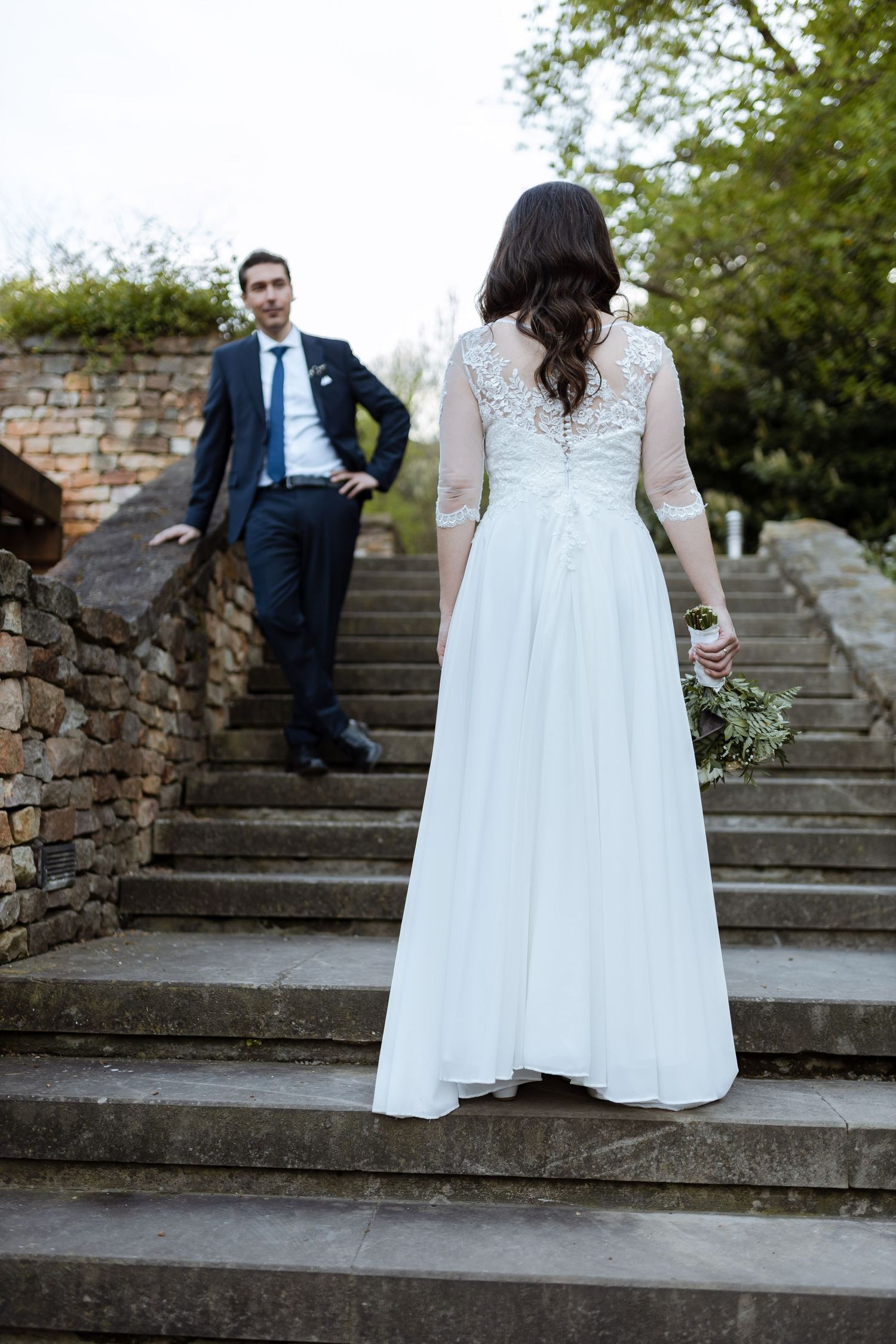 A bride in a white lace dress stands on stone steps while a groom in a navy suit leans against a brick wall.