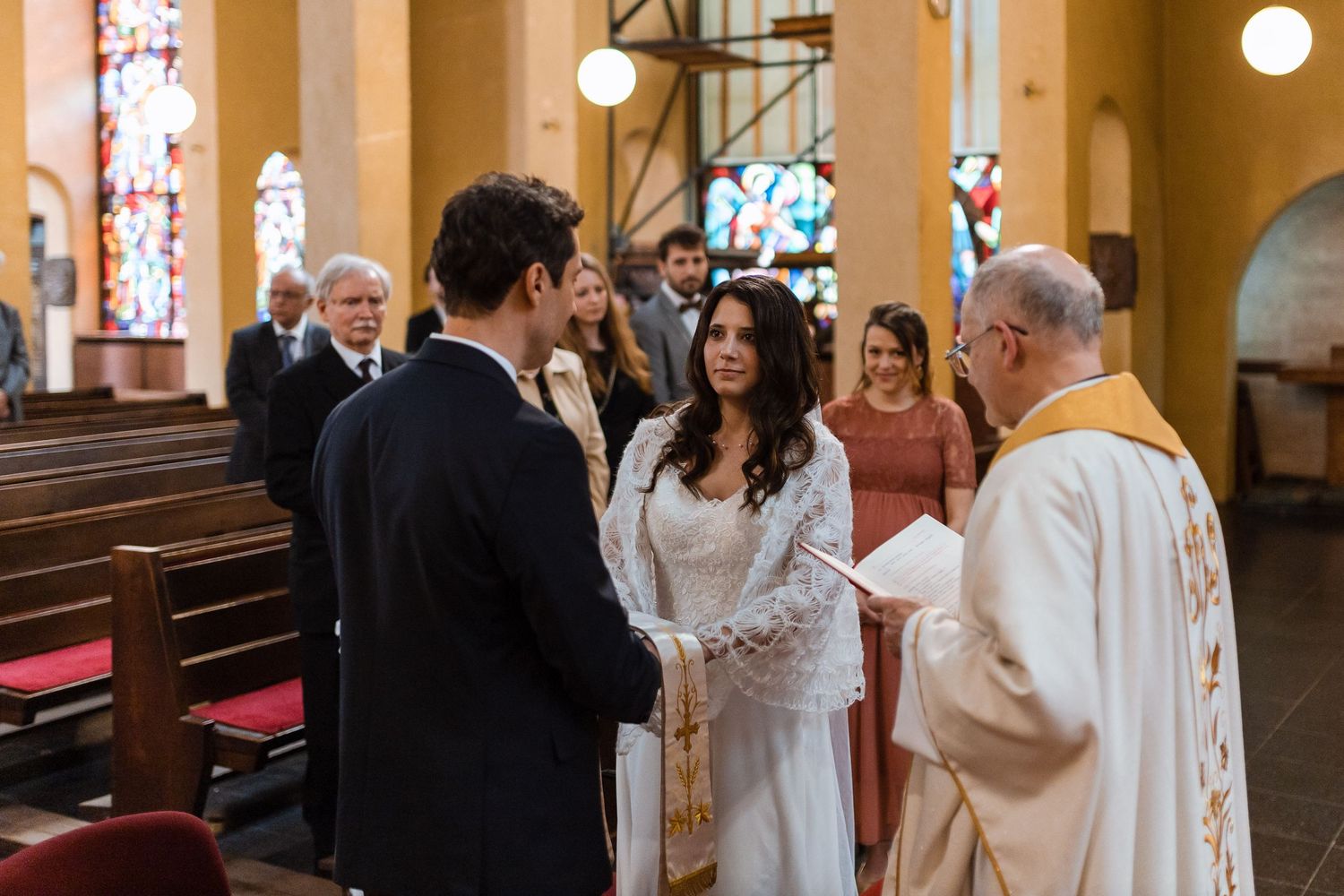 Wedding party stands in church aisle during traditional Catholic ceremony with stained glass windows in background.