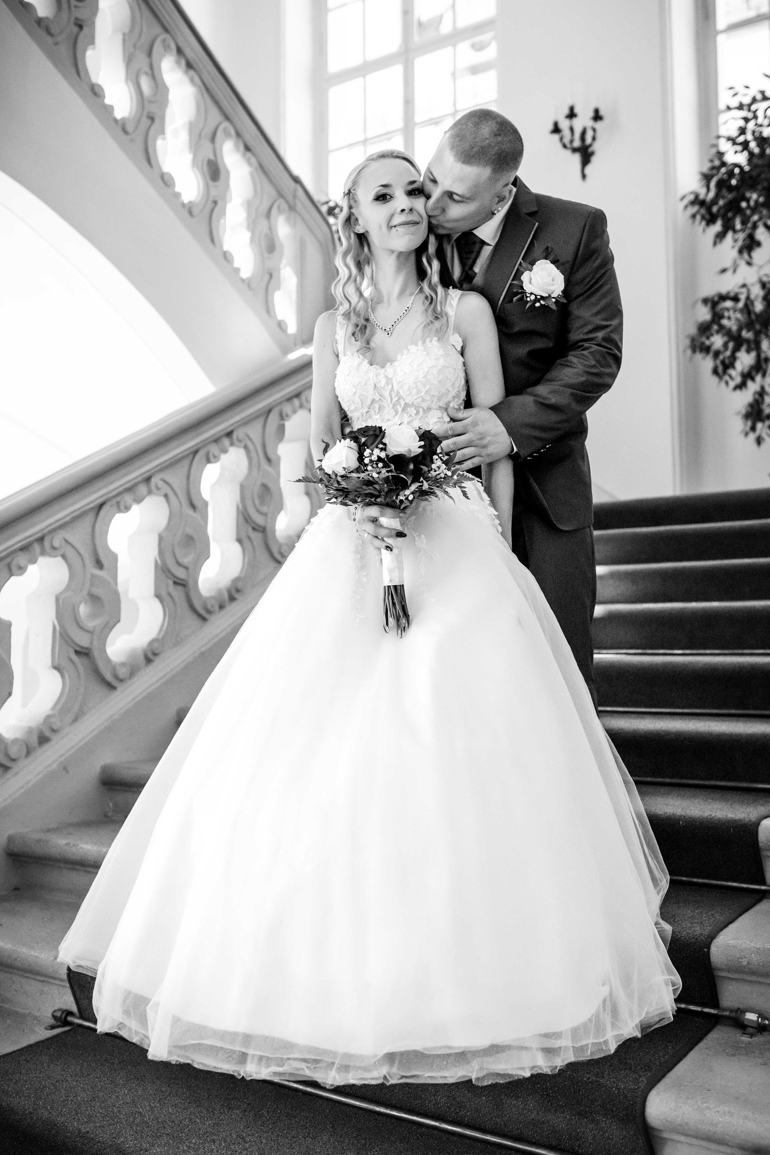 Black and white wedding photograph on an elegant staircase with ornate white railings and a bouquet of flowers.