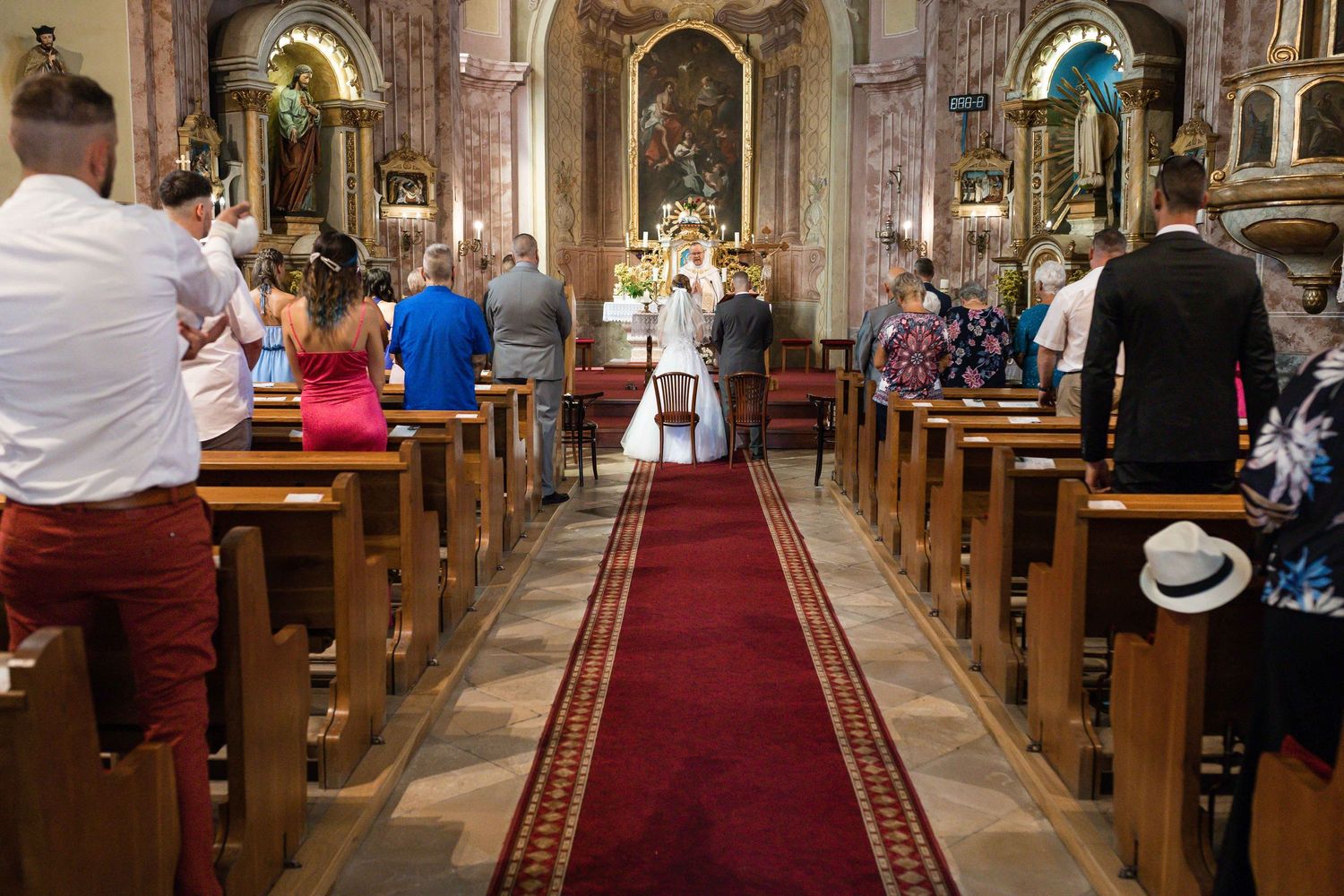 Church interior with wooden pews and red carpet aisle during a wedding ceremony.