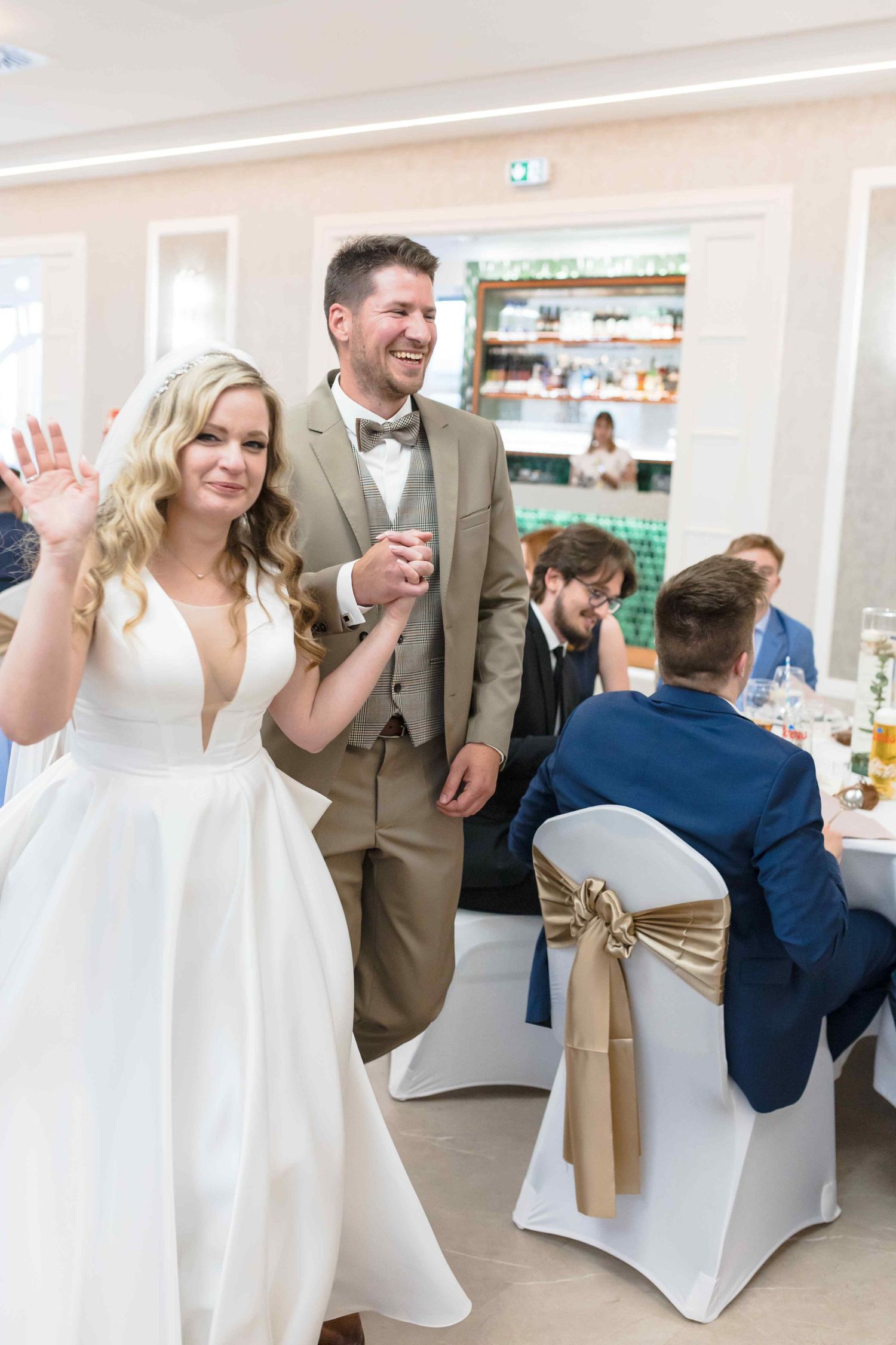 Wedding guests dancing and celebrating at a reception venue decorated with white chair covers and beige sashes.