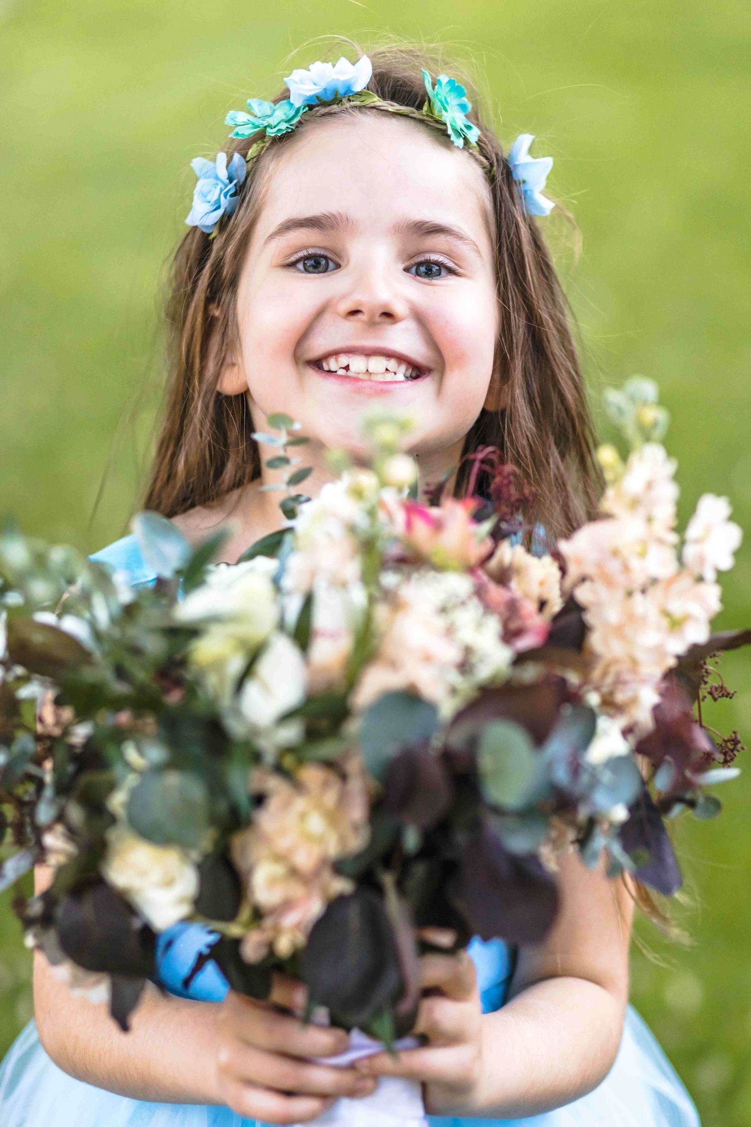 A child in a blue dress holds a beautiful bouquet of pink and white flowers against a green outdoor background.