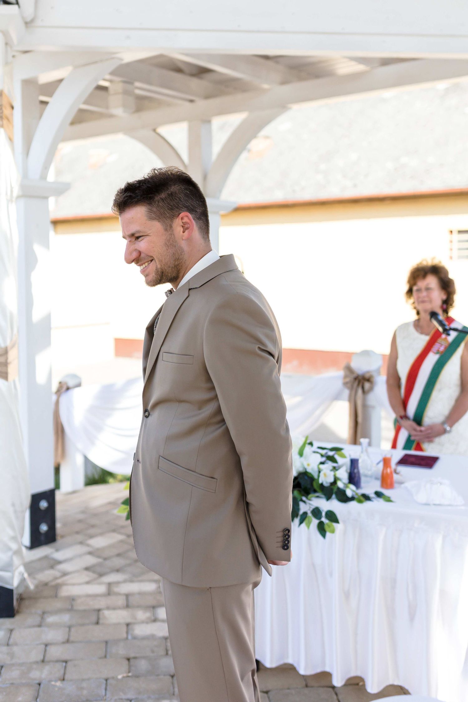 A person in a beige suit stands at a white stone wall decorated with white fabric draping at an outdoor venue.