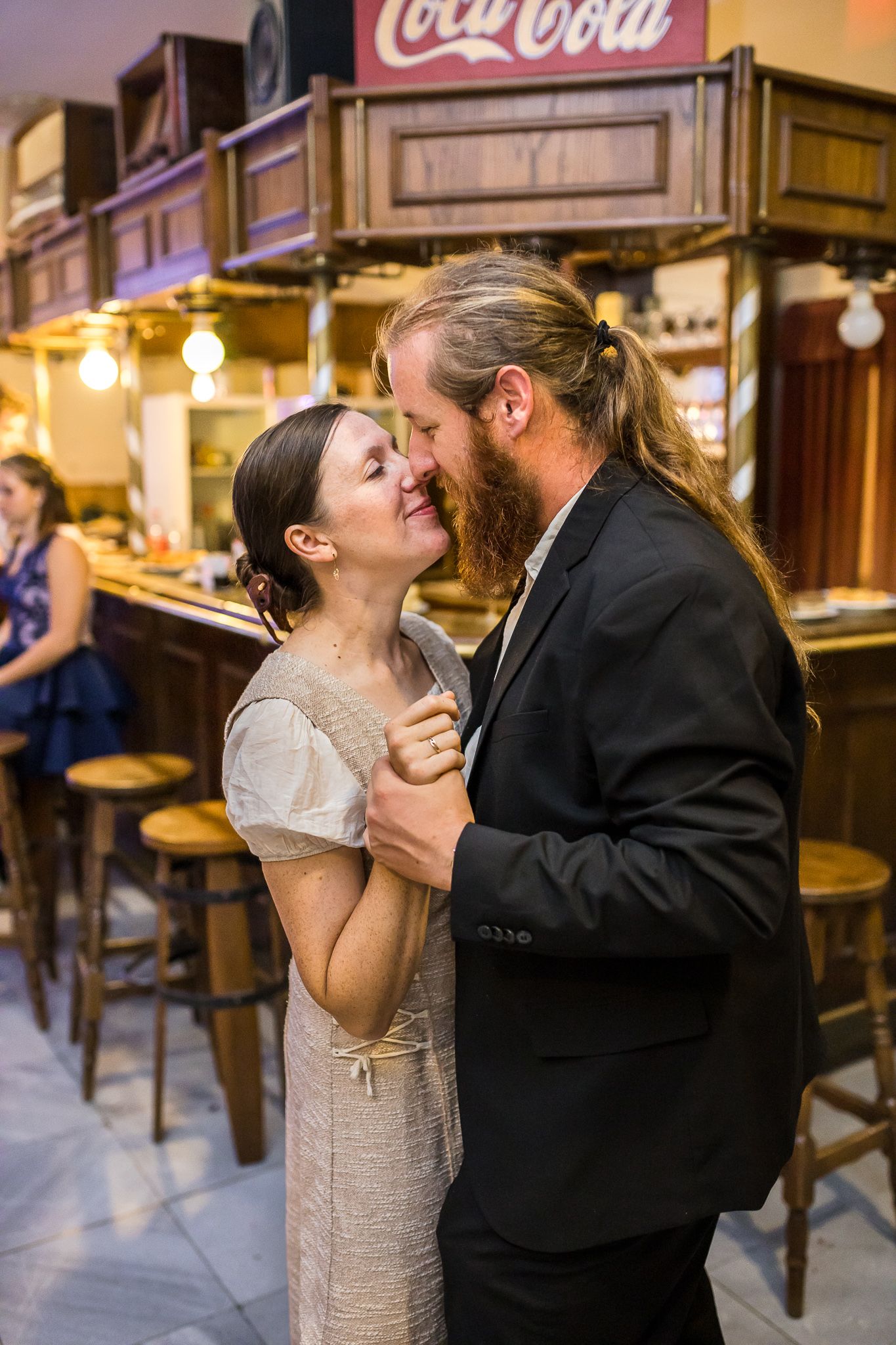 A couple shares a romantic kiss at a cozy cafe with a Coca-Cola sign visible in the background.